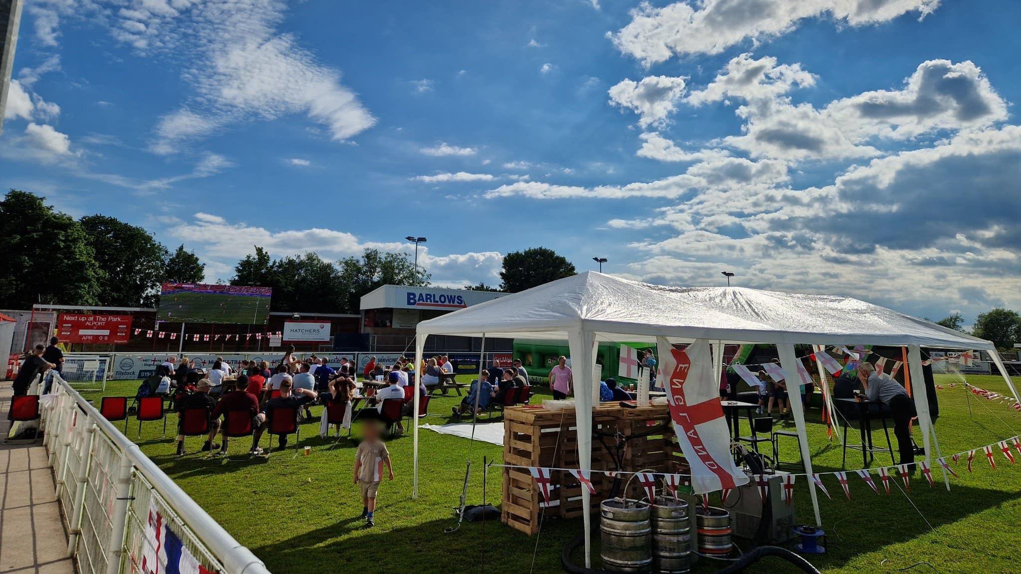 the club's pitch with people watching football on a screen