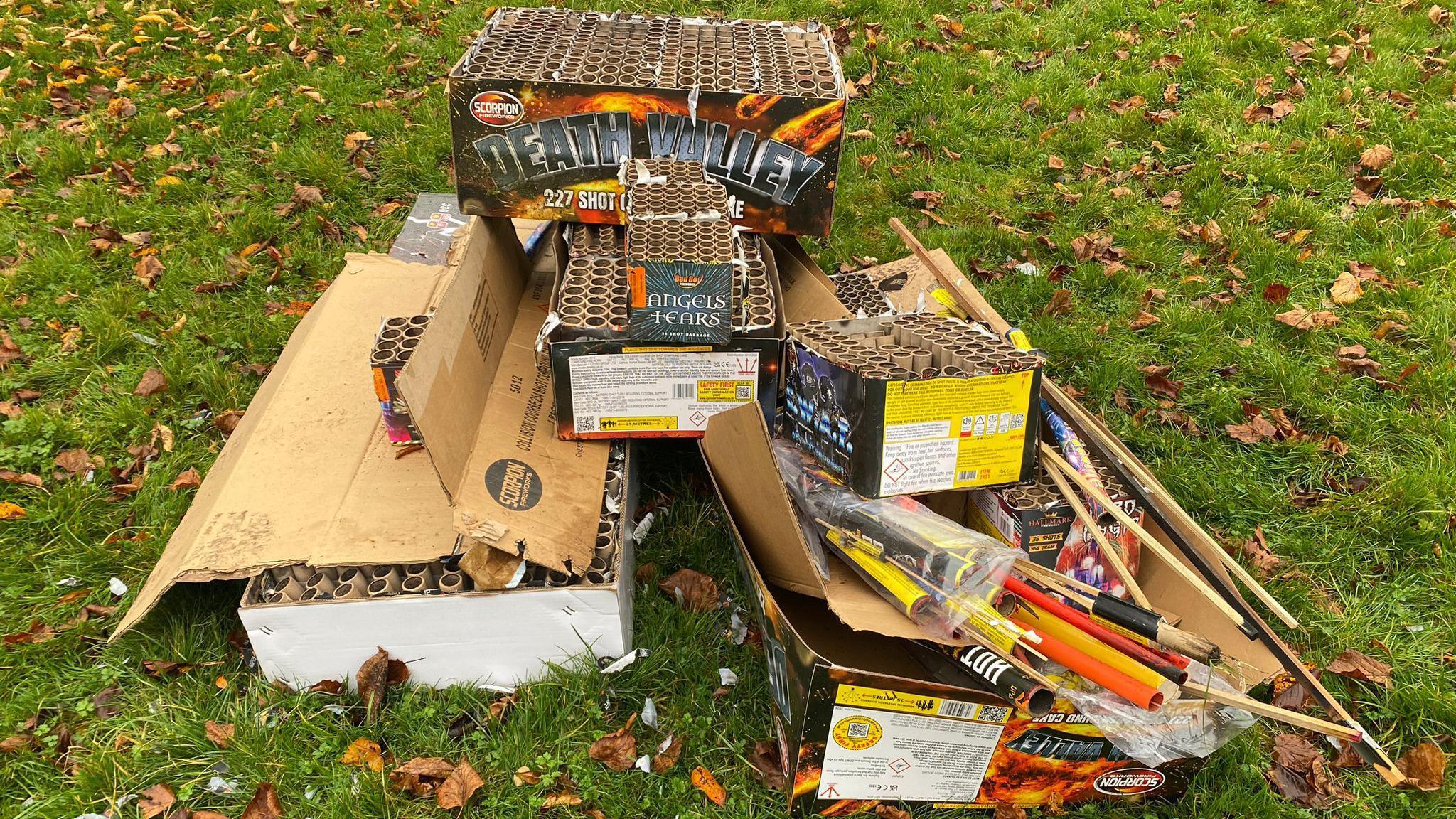 Boxes of used fireworks are left on grass and fallen leaves in a park.