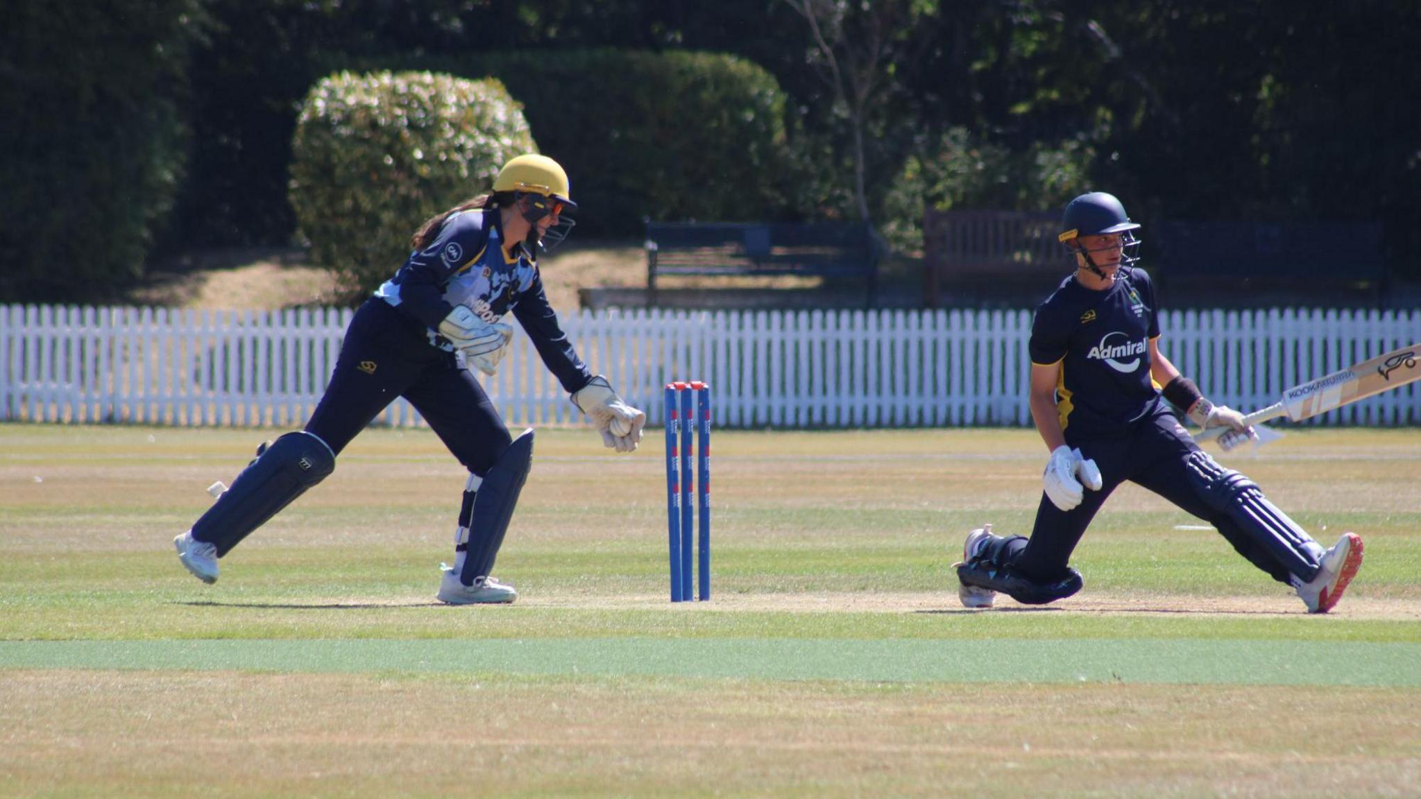 Libby, with a long brown ponytail under her yellow helmet takes the ball in her left glove as she prepares to whip off the bails 