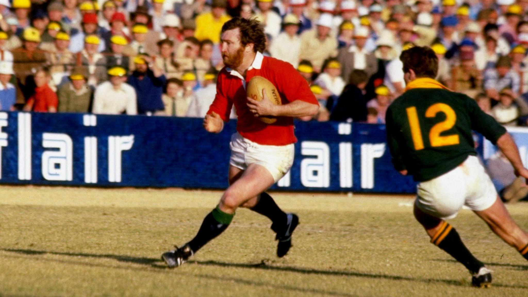 The bearded figure of Ray Gravell carries the ball in the red Lions jersey as he runs passed a South Africa player