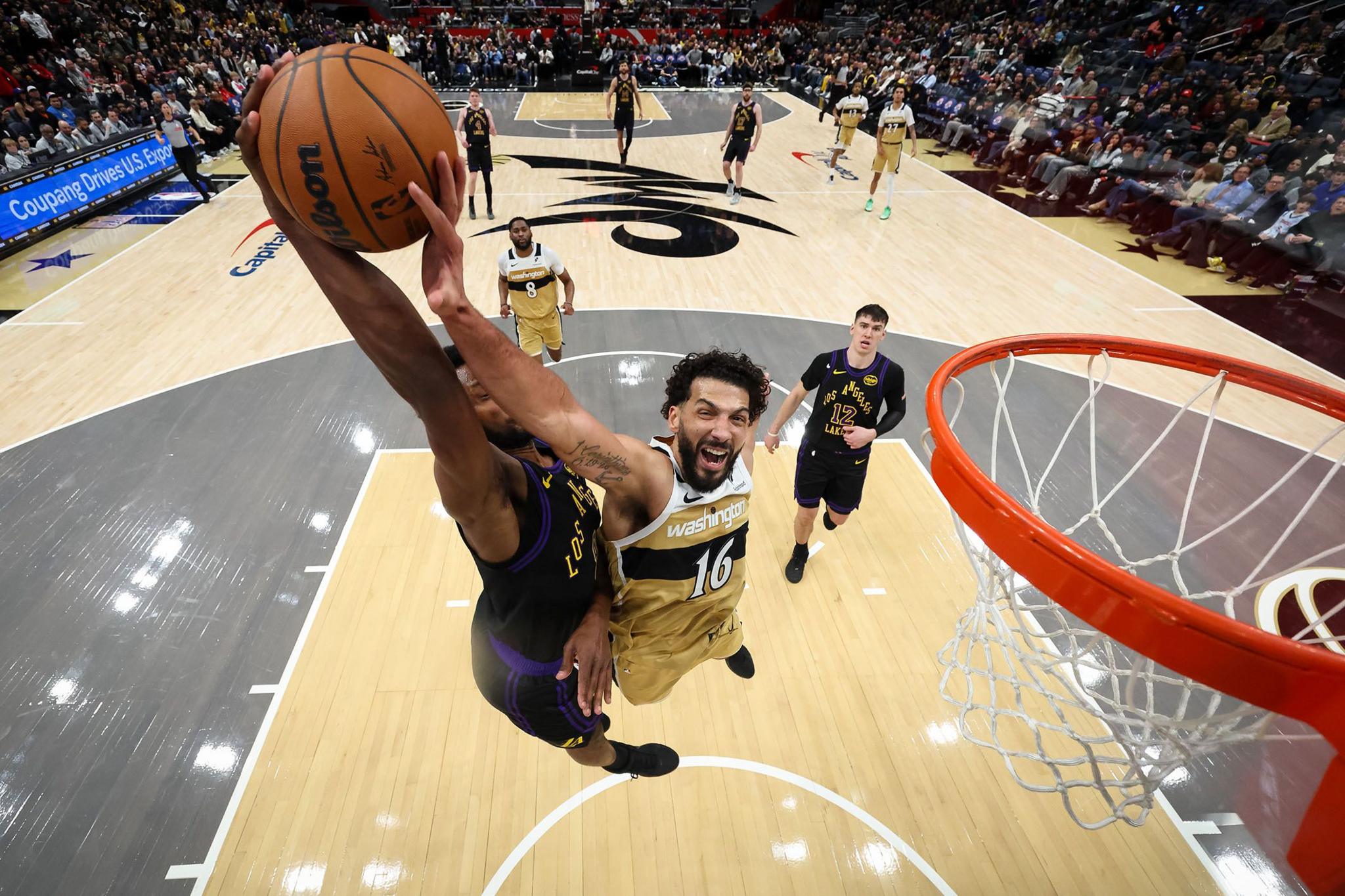 A basketball player in a purple jersey leaps toward the hoop for a powerful dunk while a defender in a gold uniform jumps to contest the shot. The action is captured from above the rim, with players and spectators visible across the court in the background.