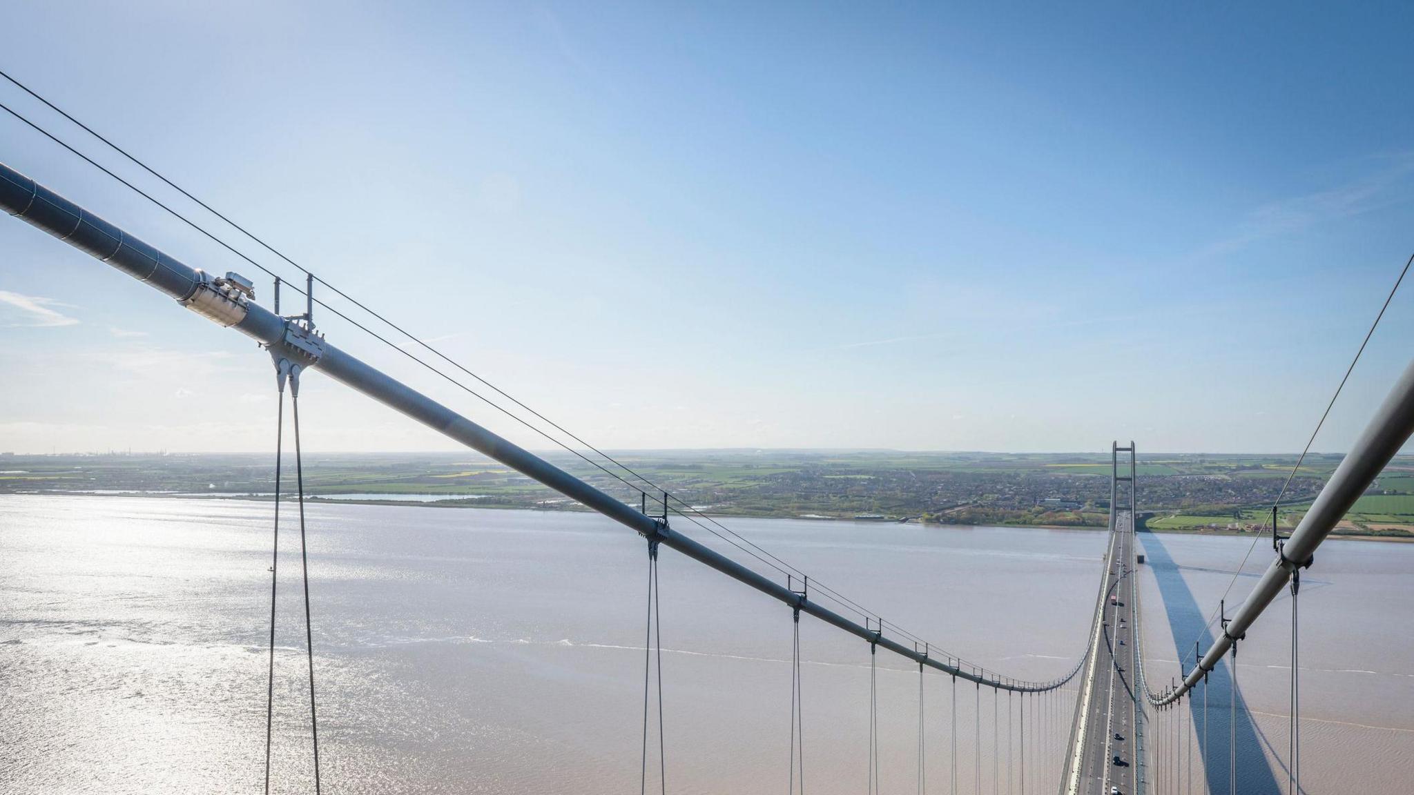 A view of the Humber Bridge over the estuary, taken from the top of the suspension bridge looking towards the town of Barton. It is a sunny day with blue skies.