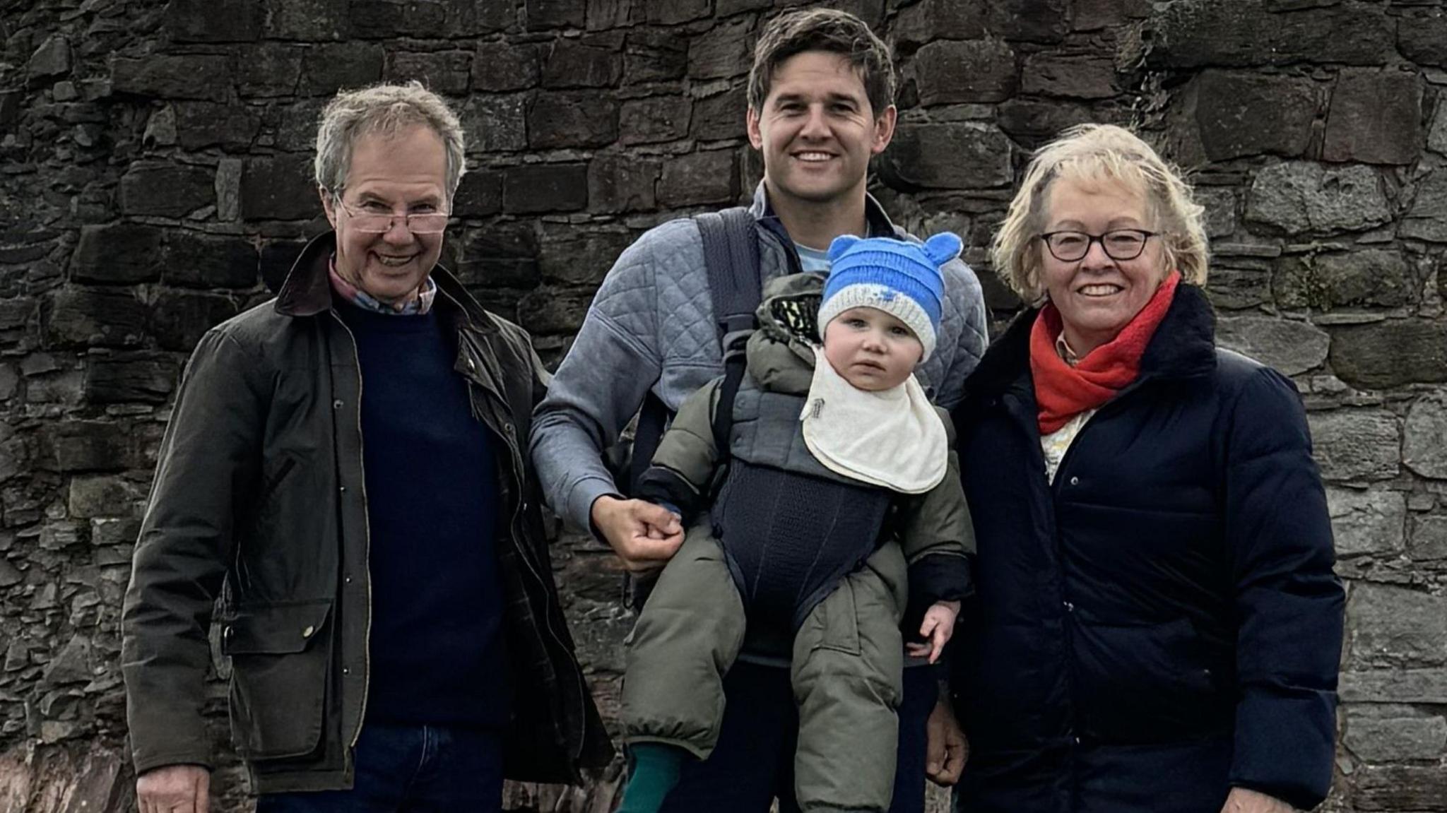 A family group standing outdoors in front of the remains of an old stone wall, part of an historic ruin or castle. The people are dressed in outdoor clothing, including jackets and boots, and the man in the centre is carrying a small child. Behind the group there is a scenic view of green fields stretching into the distance under a partly cloudy sky.