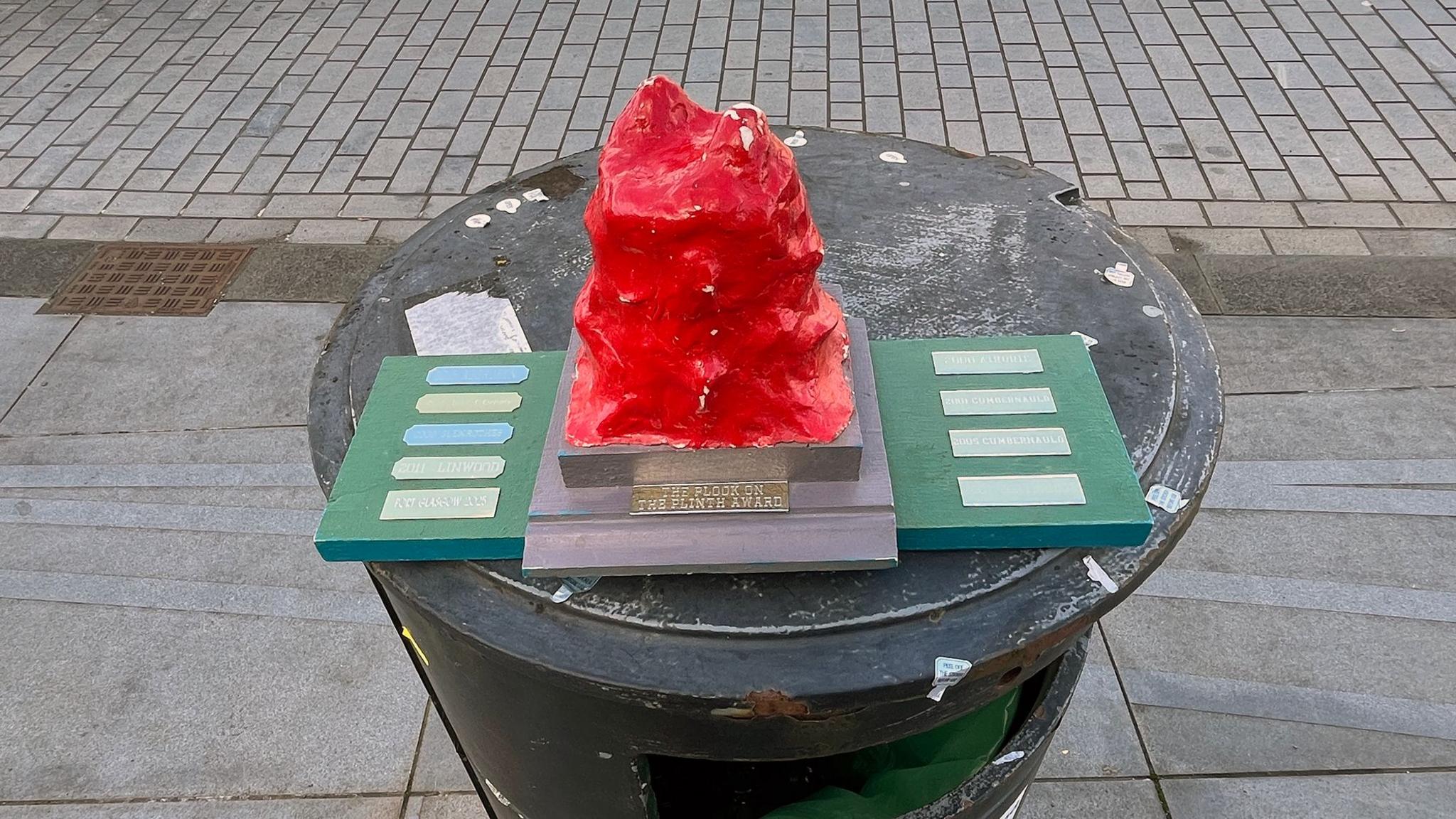 A large red pimple, shaped out of Play-Doh, on a board with small engraved metal plates showing the names of previous winners