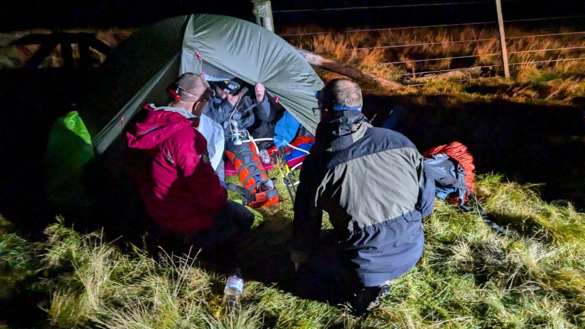 A rescue team gathers round an injured man in a small tent on some rough grassy ground near the top of a hill