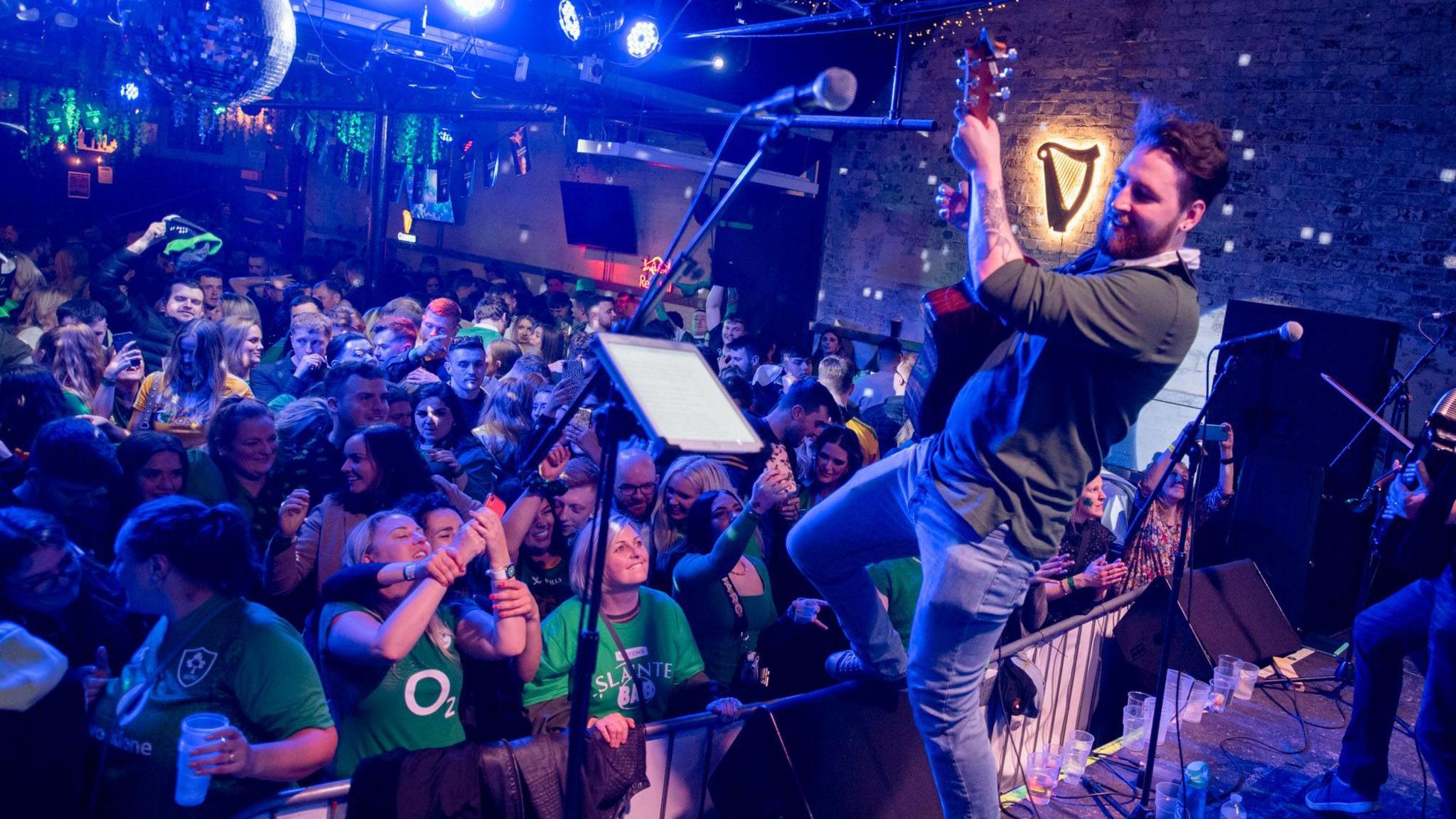 A man on stage is playing a guitar whilst raising one foot onto a barrier separating him from a crowd inside a night club. The room is lit in a blue light and there are microphones and disco balls visible.