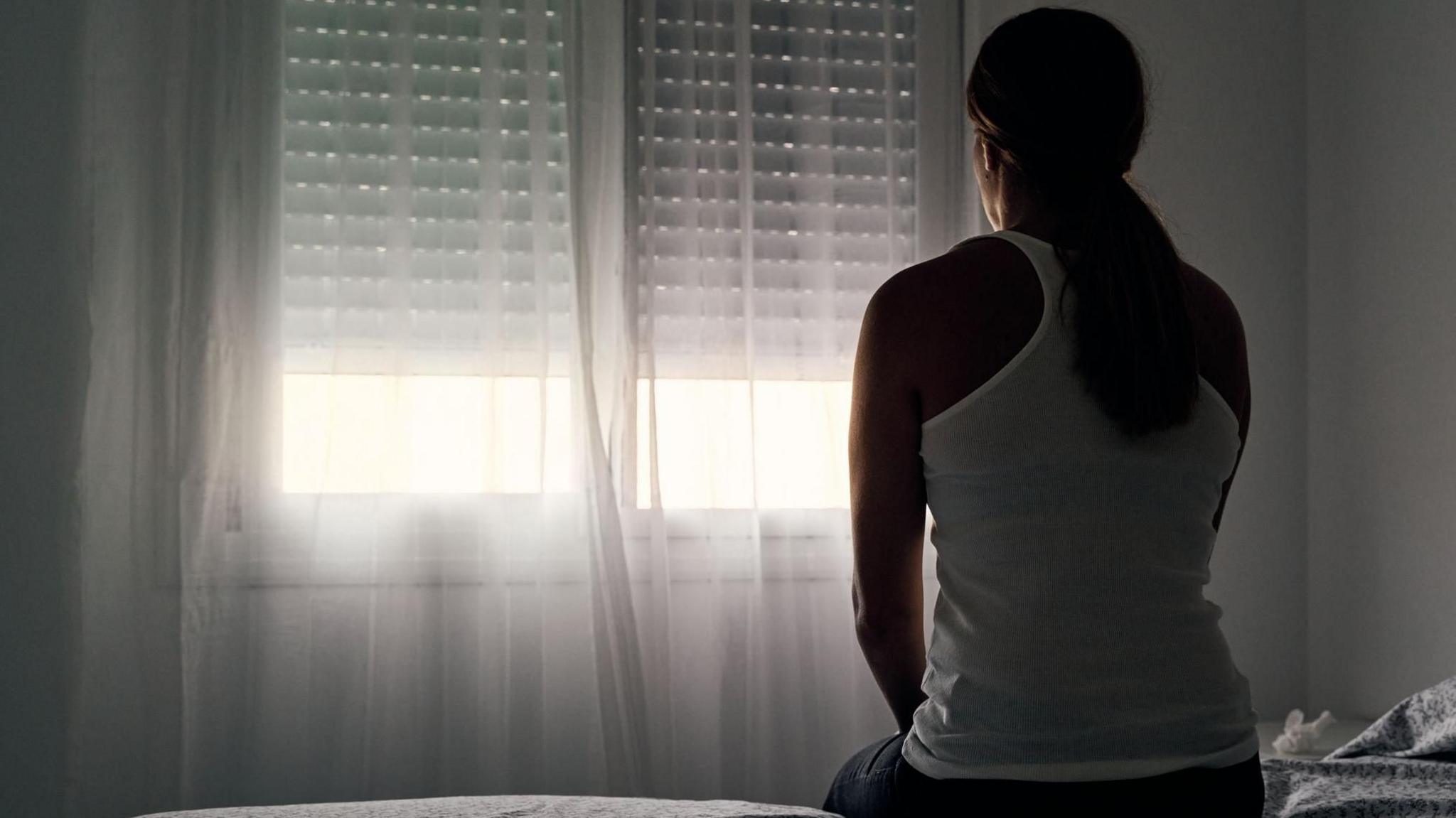 A stock image of a woman sitting on a bed staring out of a window covered by a net curtain.