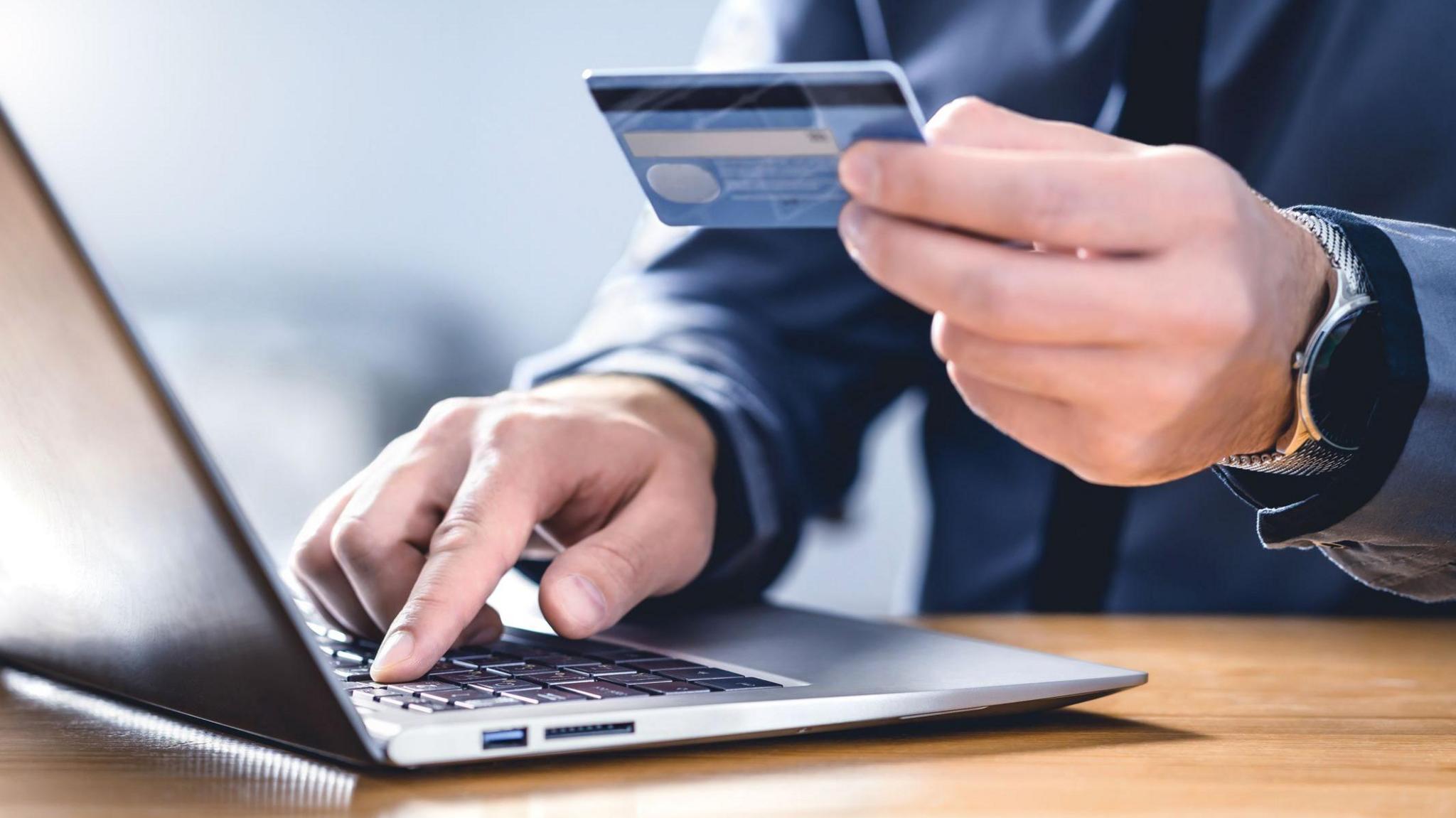 Close up of a man holding a bank card in one hand and tapping on a laptop keyboard with the other.