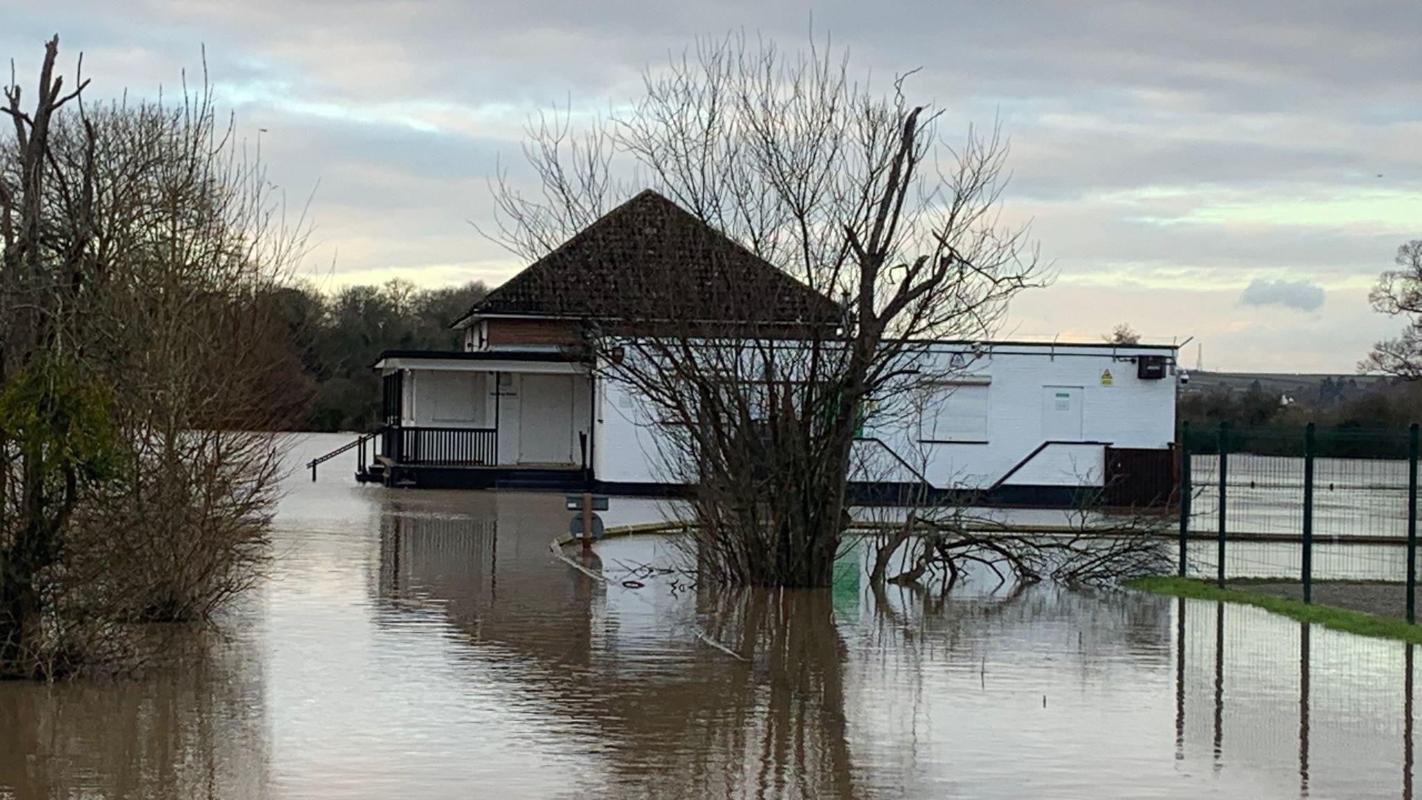 A single-story white building with a black roof sits in the middle of flood water. Several trees can also be seen emerging from the water.