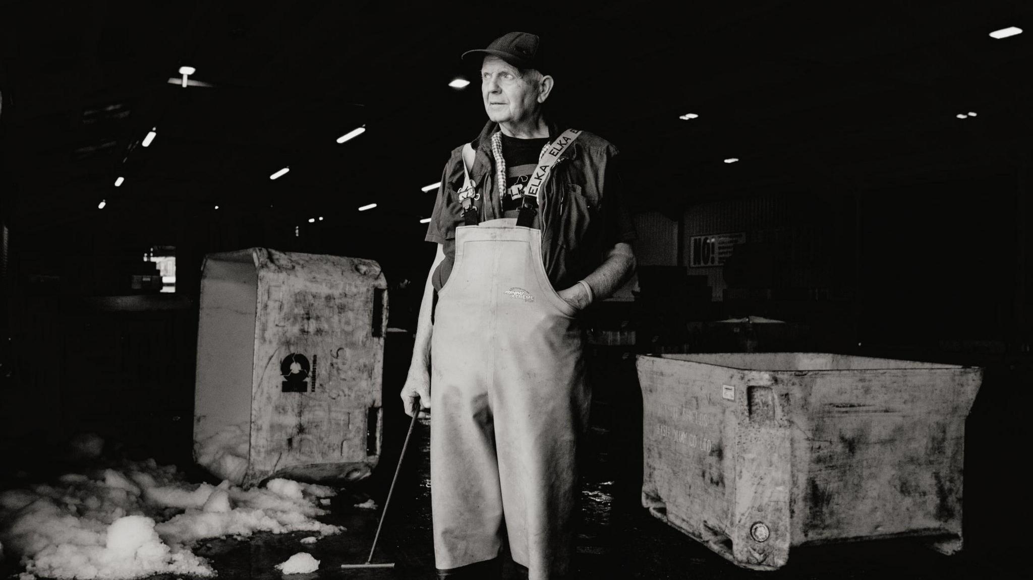 A black and white image of an older fisherman in the fish hall in North Shields. Ice used to store fish can be seen melting on the ground behind him. He is wearing waterproof bib and braces. 