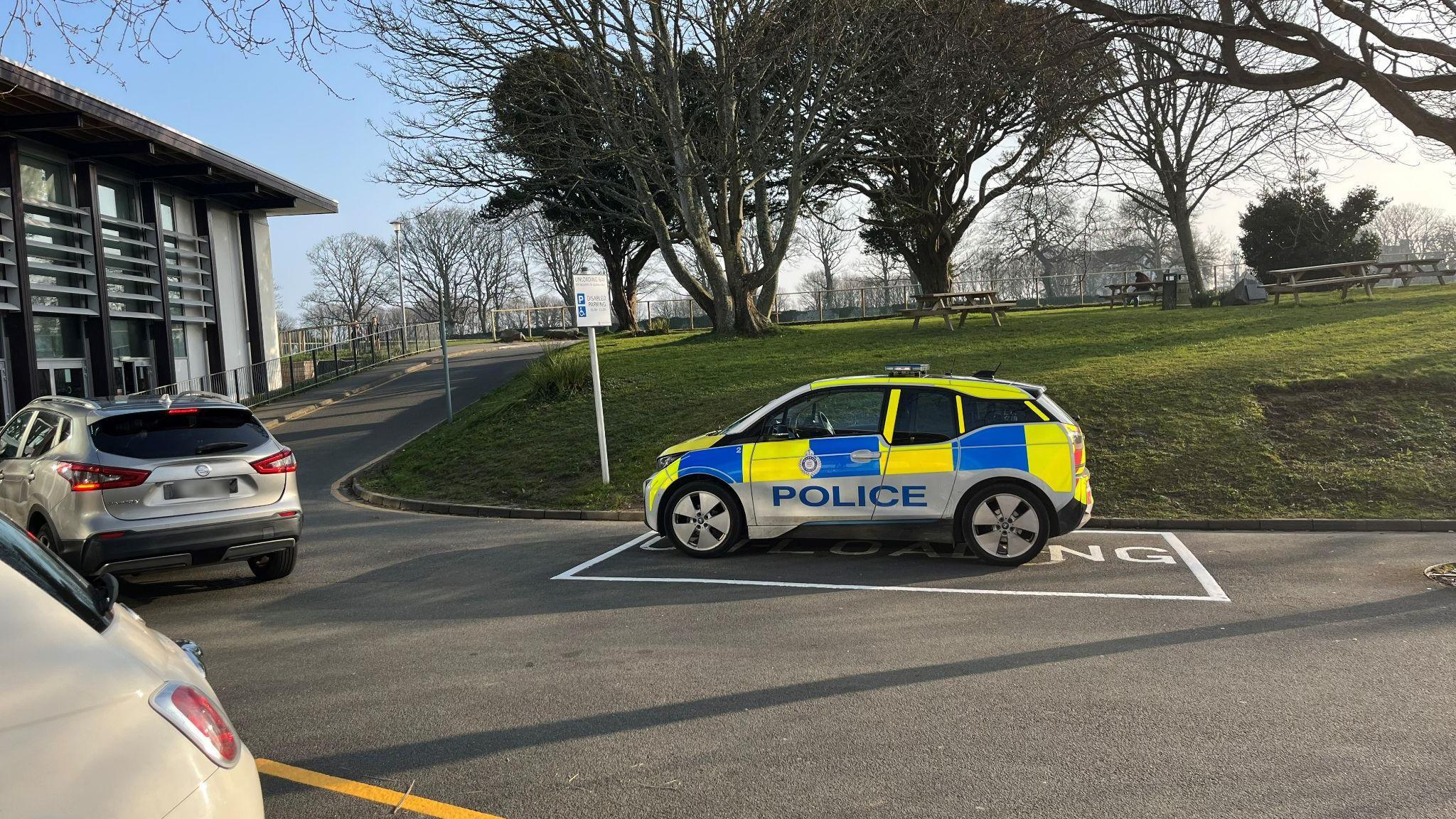 A police car parked outside Beau Sejour Leisure Centre in Guernsey on a sunny day. A silver SUV is driving past the police car.