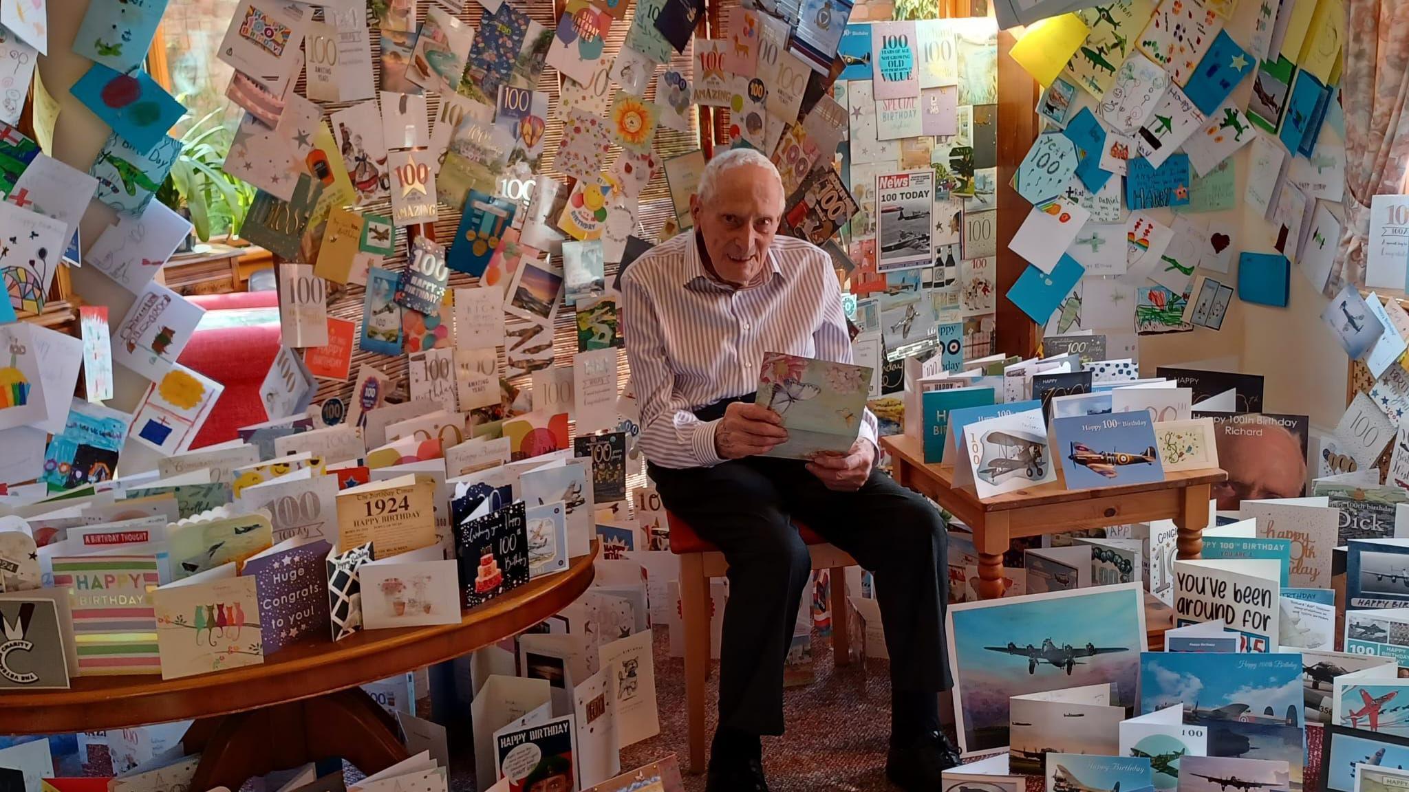 An old man in a white shirt and black trousers holds a card and smiles at the camera, in a room that is filled with birthday cards - on the floor, on tables, and covering every inch of the walls