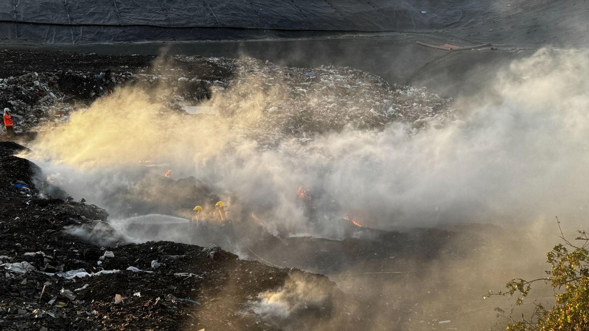 A wider view of the smoke billowing from the landfill site. Two firefighters hose down part of the landfill site.