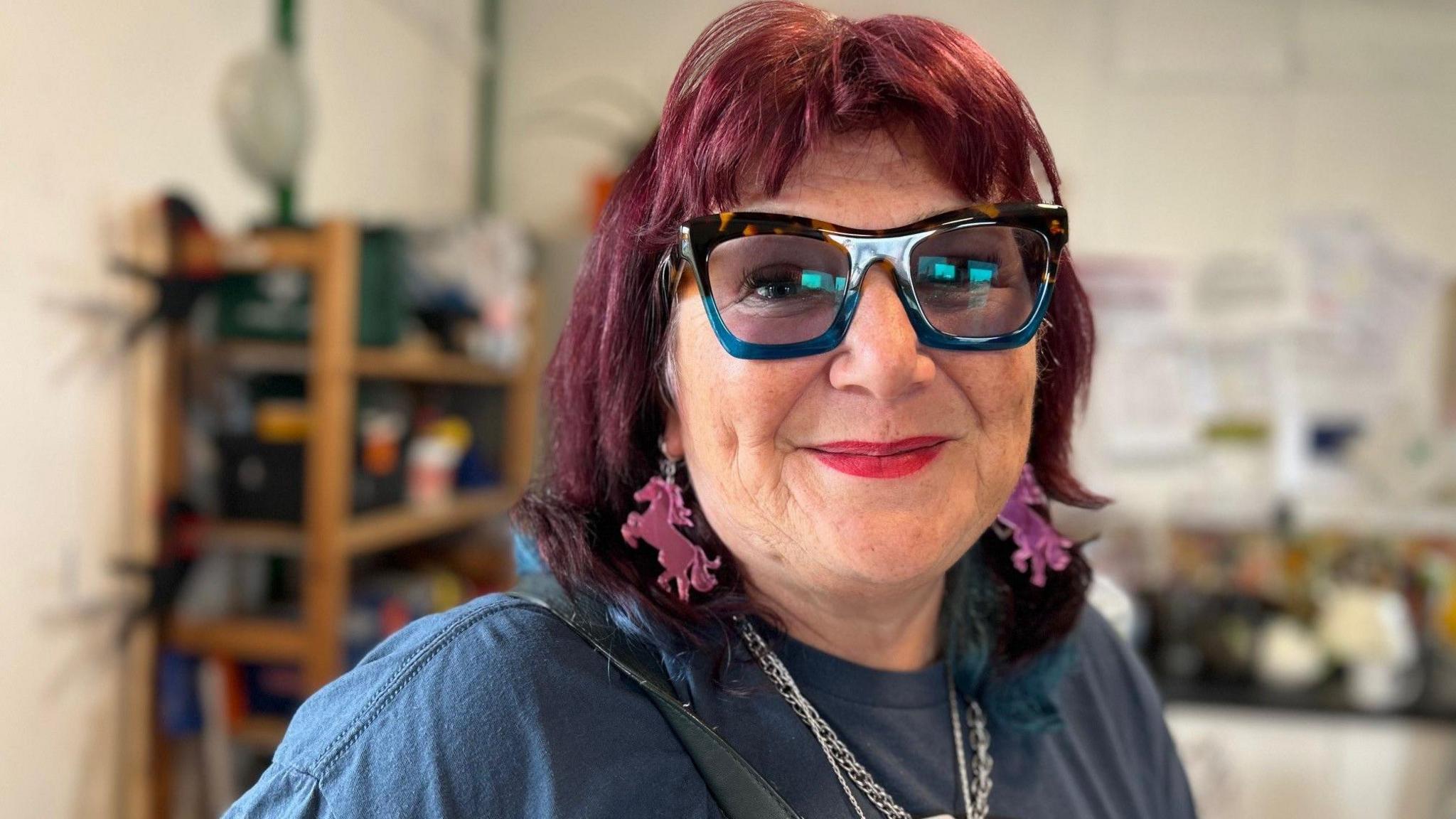 Susan has red shoulder length hair and is wearing thick-rimmed blue and leopard-print glasses. She has pink unicorn-shaped earrings and is wearing a blue t shirt. 