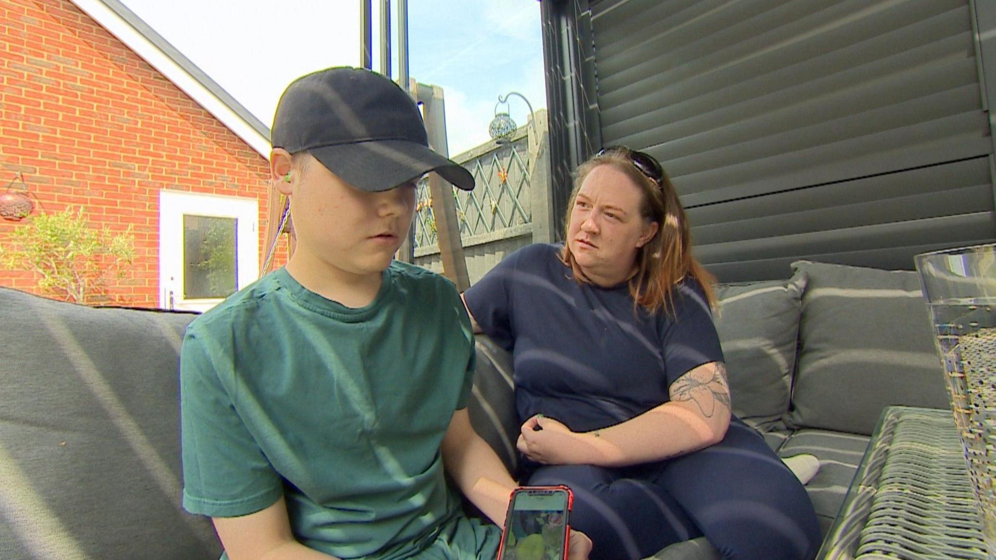 Tora sitting on an outdoor sofa in the garden with her son Mason. Mason is wearing a black baseball cap and green t-shirt and is holding a phone. Tora is wearing blue and is looking at Mason.