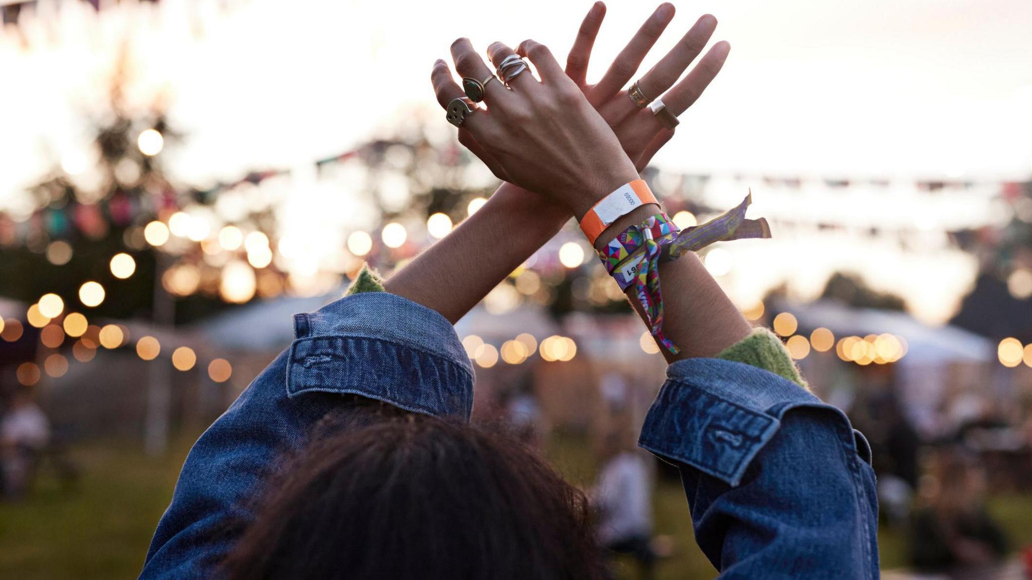 A woman with brown hair is wearing a denim jacket. She is at a festival with fairy lights and tents blurred in front of her. Her arms are in the air as if she is dancing. She has wristbands on her wrist and rings on her fingers