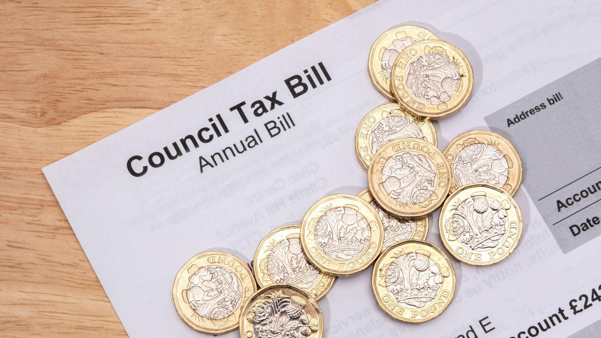 A white piece of paper that reads "council tax bill, annual bill". There are twelve £1 coins sitting on top of it. The items are on a light wood table