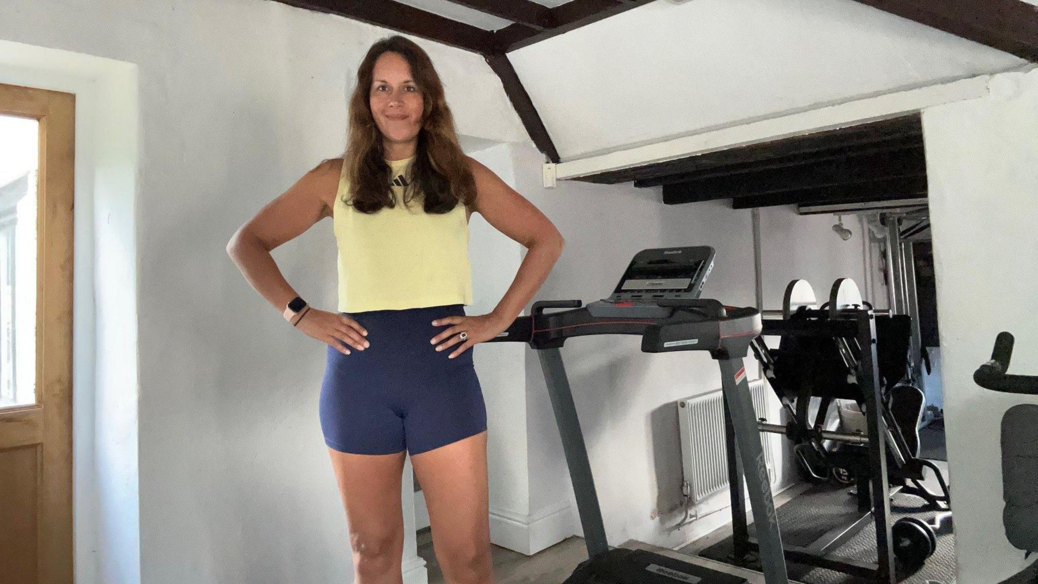 Emily Brady, with her hands on her hips, wearing a yellow tank top and blue cycle shorts, stands in front of a home gym. 