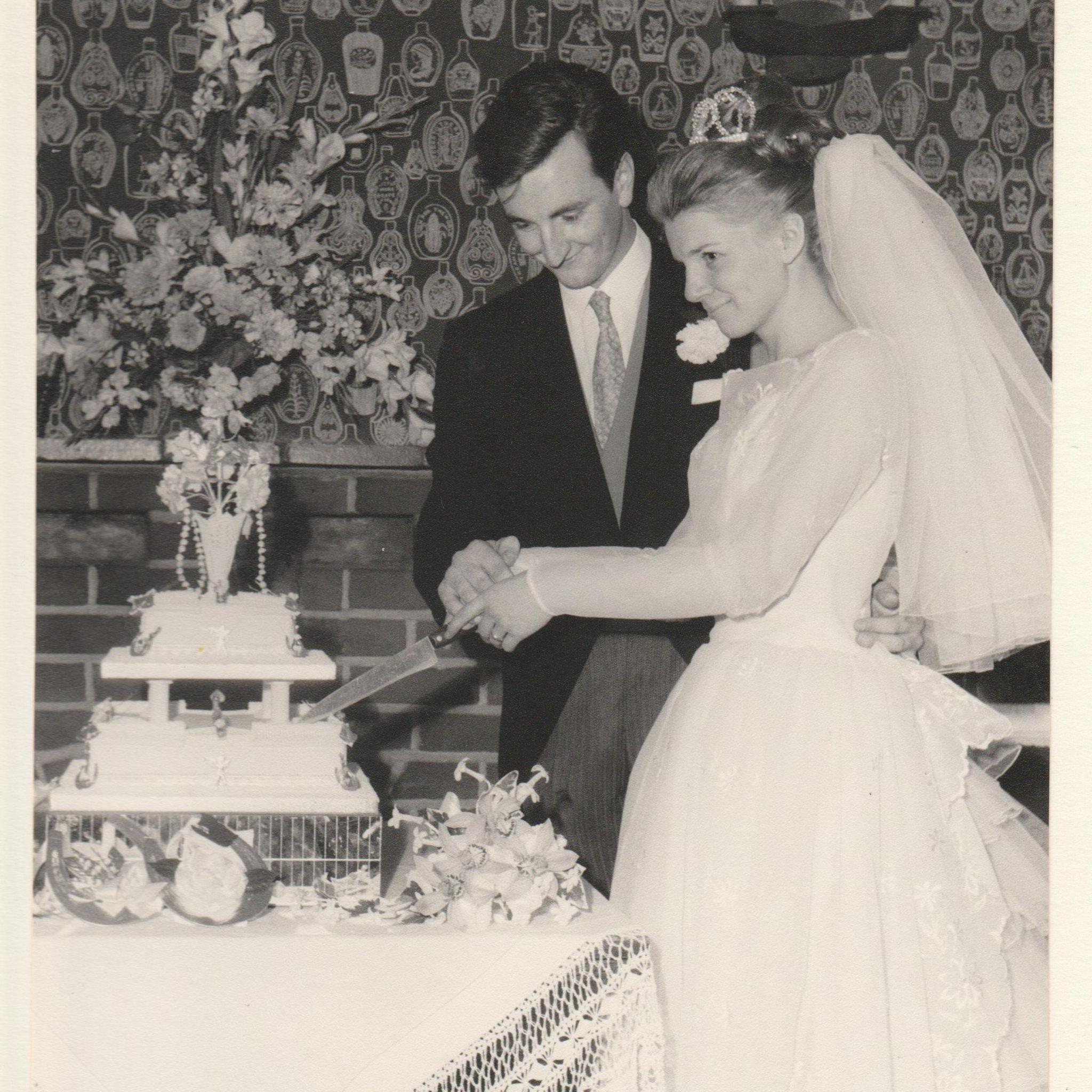 A bride and groom on their wedding day both holding a knife as they cut their wedding cake, a two-tiered cake placed on a table with a bouquet of flowers on top.