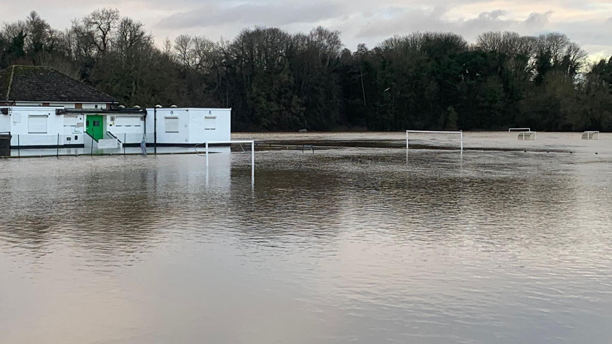 Football goalposts sit in the middle of flood water. A white building with a black roof is inaccessible due to the water.