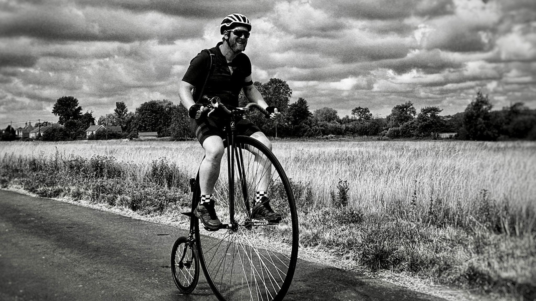 Richie Mason in a black and white image. He is riding a penny farthing on what appears to be a single track road in a field.