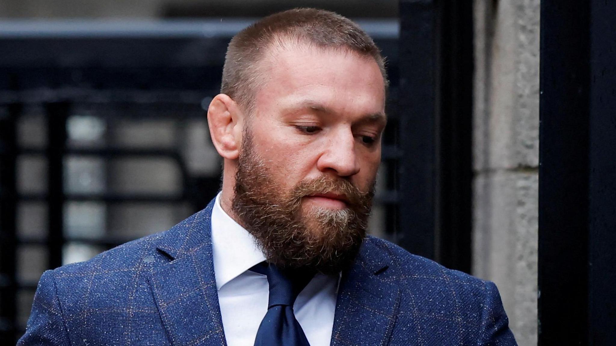 Conor McGregor, who has short, light brown hair and a thick brown beard and wears a blue-checked suit jacket over a white shirt and a navy tie.