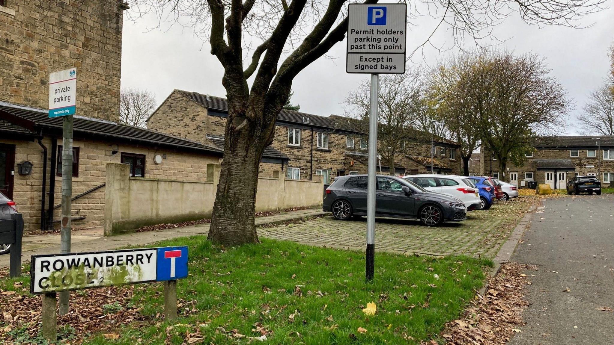 A white street name sign with black lettering and a symbol showing it is a dead-end with cars parked in a row and houses in the background.  