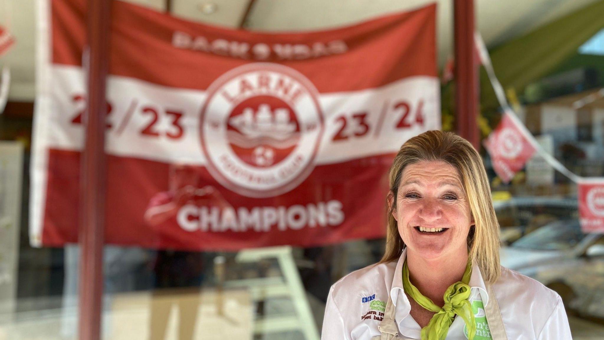 A woman in a white shirt and a beige apron stands in front of a Larne flag in a bakery window. The flag is red with a white horizontal stripe running through the middle. Written on the flag is ‘Back 2 Back Champions, 22/23, 23/24’ with the Larne crest in the middle. The Larne crest is a white circle with a red middle. In the middle is a white sail boat.
