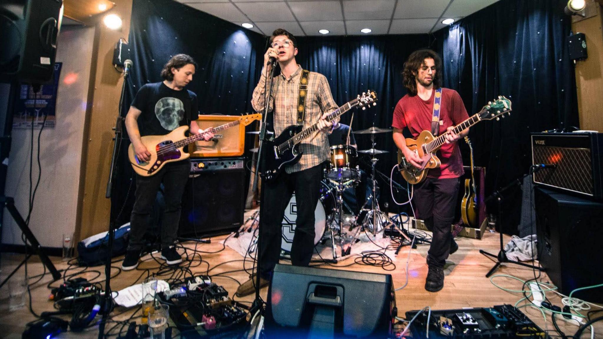 A band play on a stage surrounded by sound equipment. Three band members are standing and each holding a guitar. The background is a black curtain with lights and behind the guitarists, there is a drummer sitting at a drum kit.