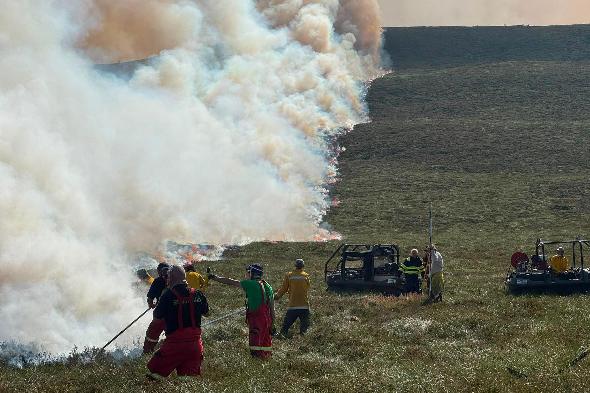 Firefighters and volunteers work to contain a large moorland wildfire as a long line of flames advances across a hillside. Thick plumes of white and brown smoke rise into the sky while crews use hoses and equipment vehicles positioned on the grassland below.