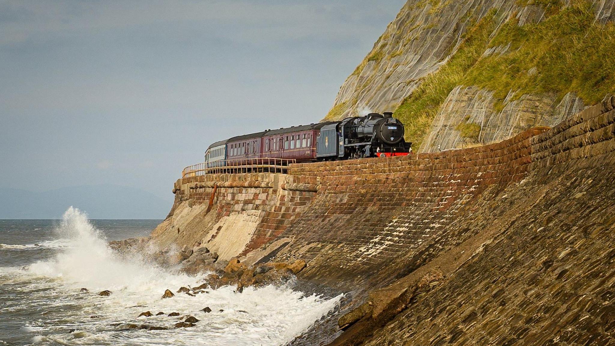 A train pulled by a steam engine runs along a track between a crashing wave in the sea and very steep cliff.