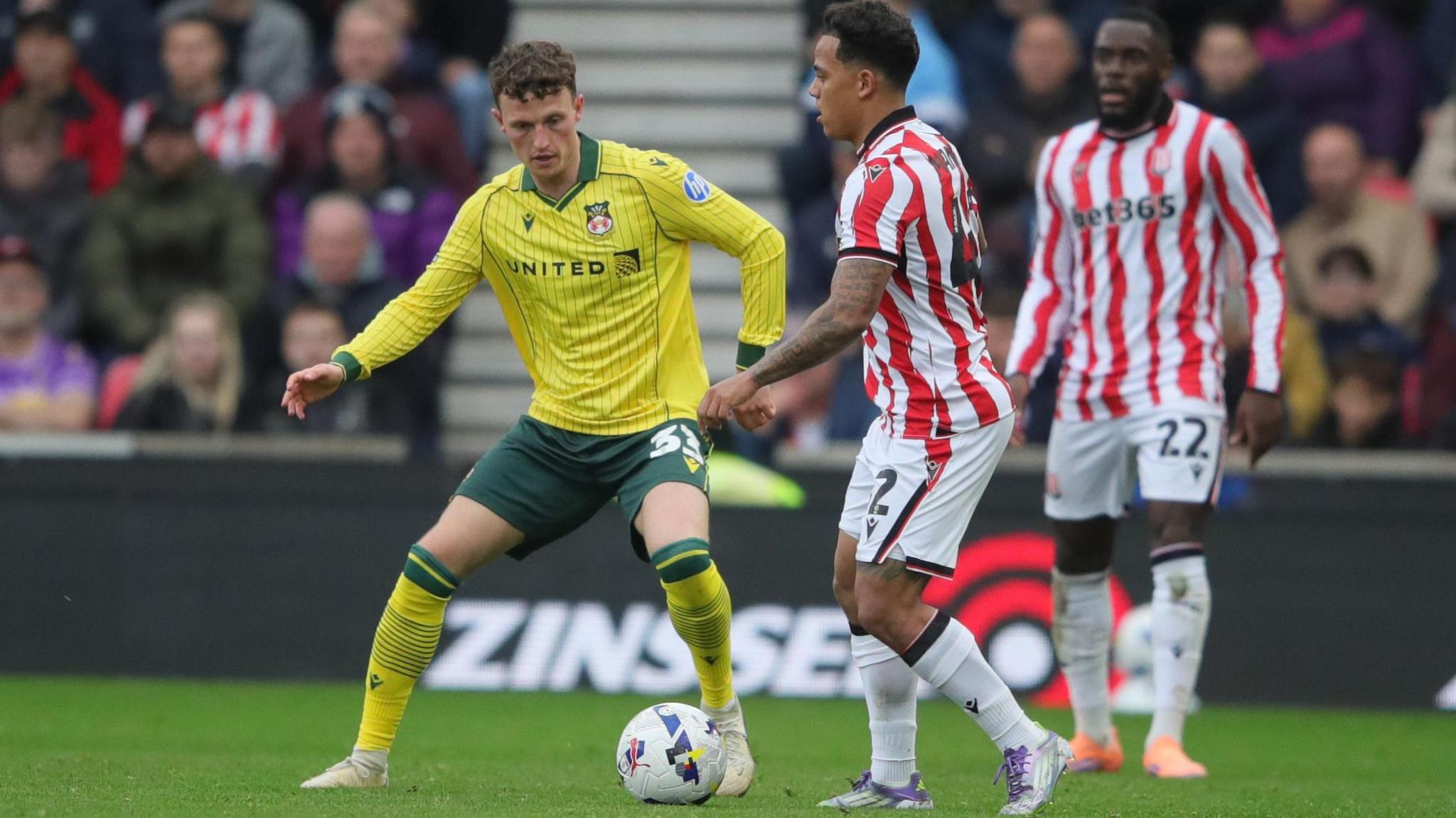Nathan Broadhead in a yellow Wrexham away shirt closes down Stoke's Milion Maenhof in possession of the ball