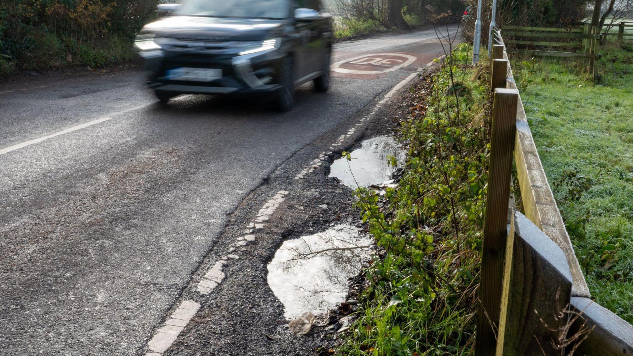 Potholes on the side of a 30mph rural road. A 4x4 is driving along towards the middle.