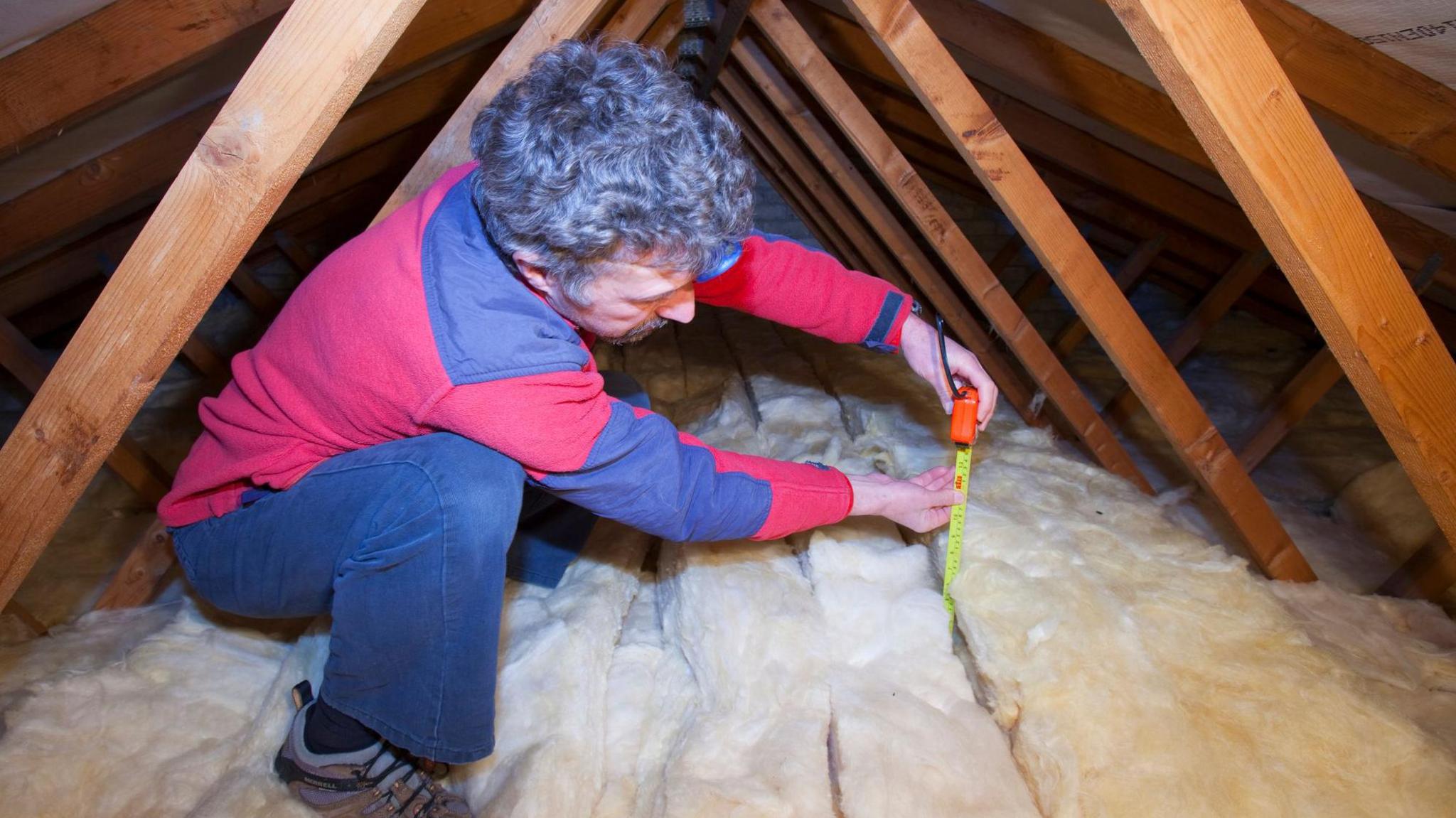 A library picture of a man measuring the depth of insulation in a house loft or roof space.