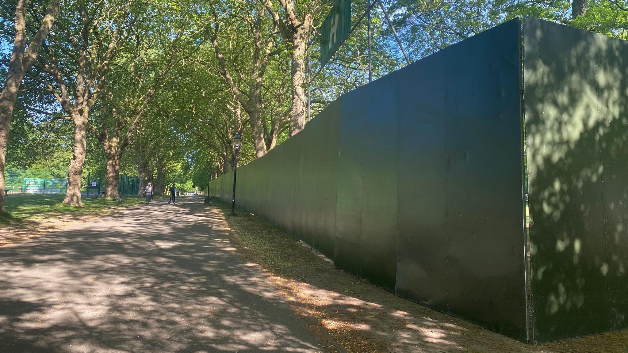 A large green fence running the length of a path in Sefton Park. People are walking in the distance.