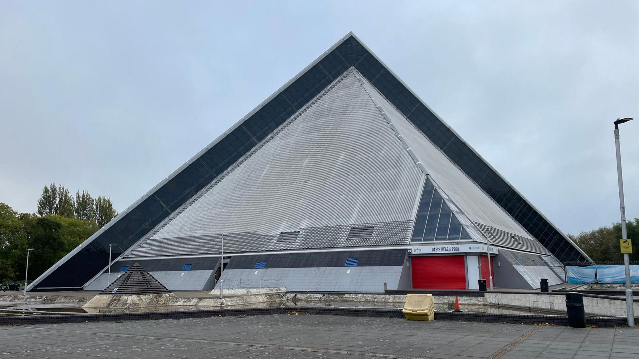 Grey pyramid building, with a red section on one corner of the base. There is a small grey barrier in front and a paved area in the foreground. There are trees behind.
