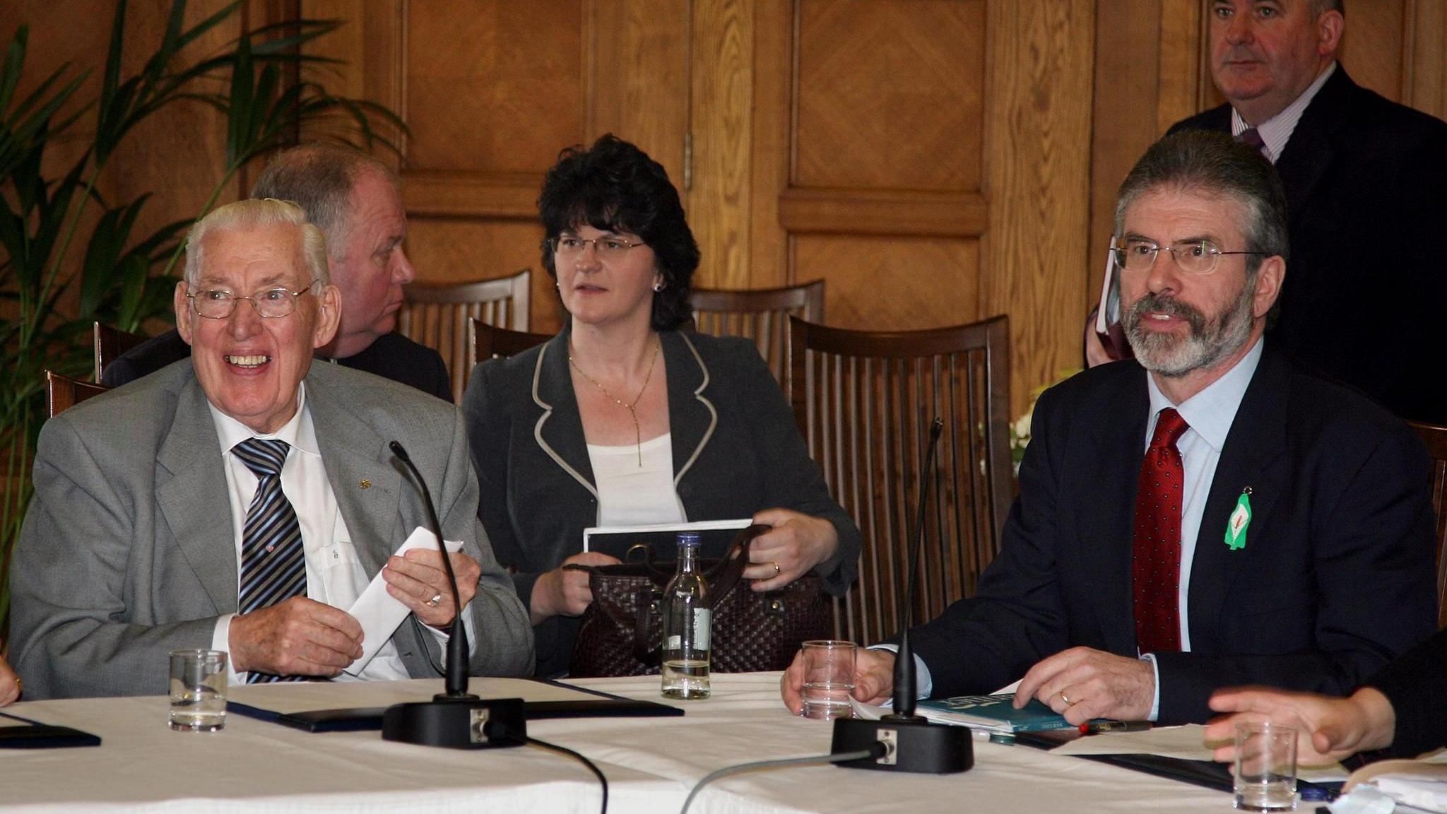 Ian Paisley and Gerry Adams sitting on two sides at the corner of a table with a white tablecloth and microphones on it.