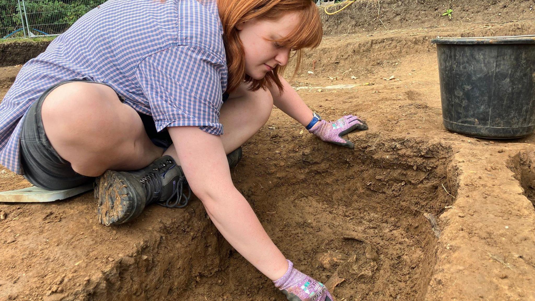 Elearnor Rawlings has ginger hair and is wearing black shorts, a blue checked shirt, and purple gloves. She's sitting cross-legged on the ground and leaning into an archaeological pit to excavate something.