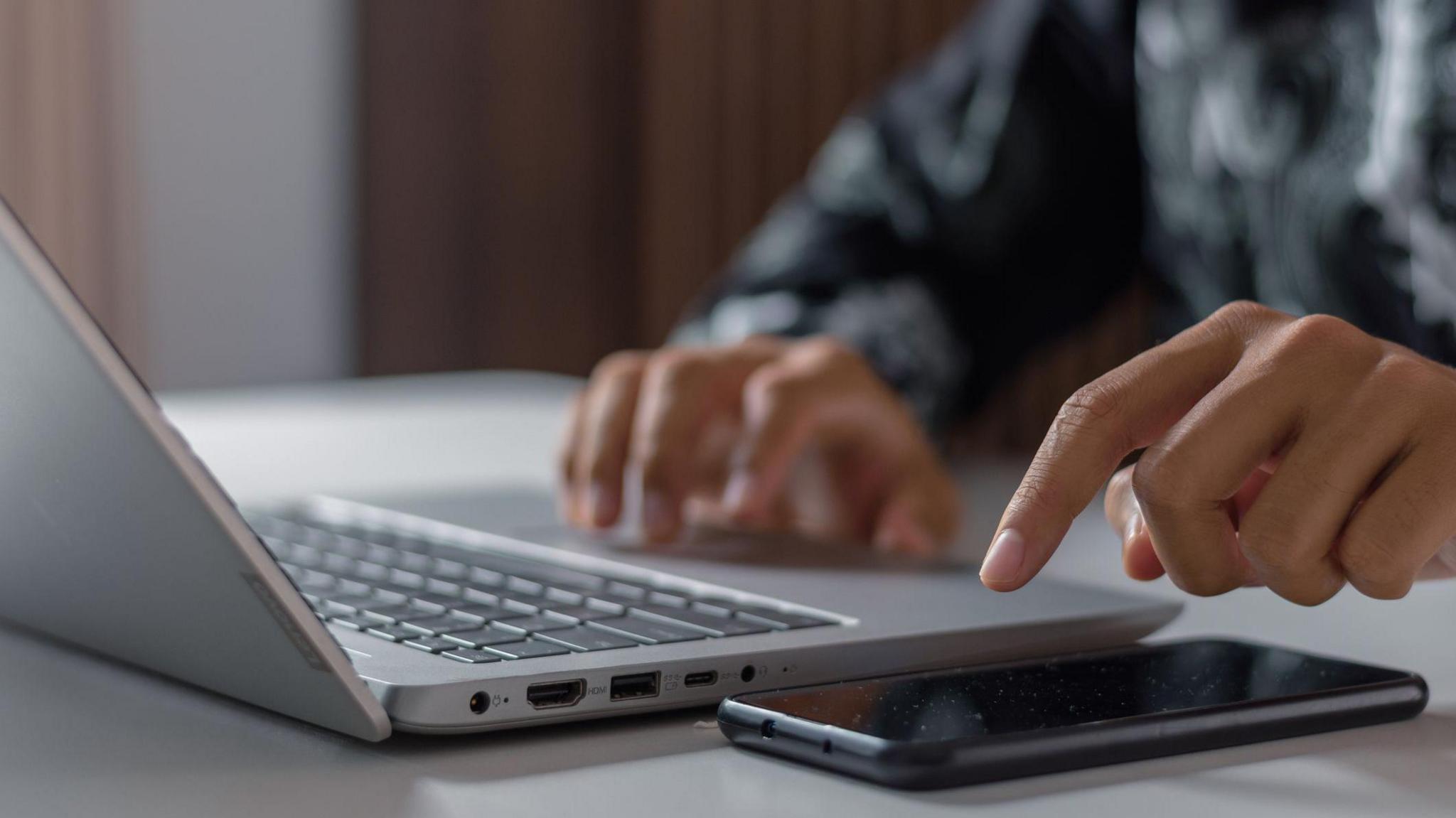 A man sits at a desk with one hand on his laptop mouse and the other hovering over his smartphone.