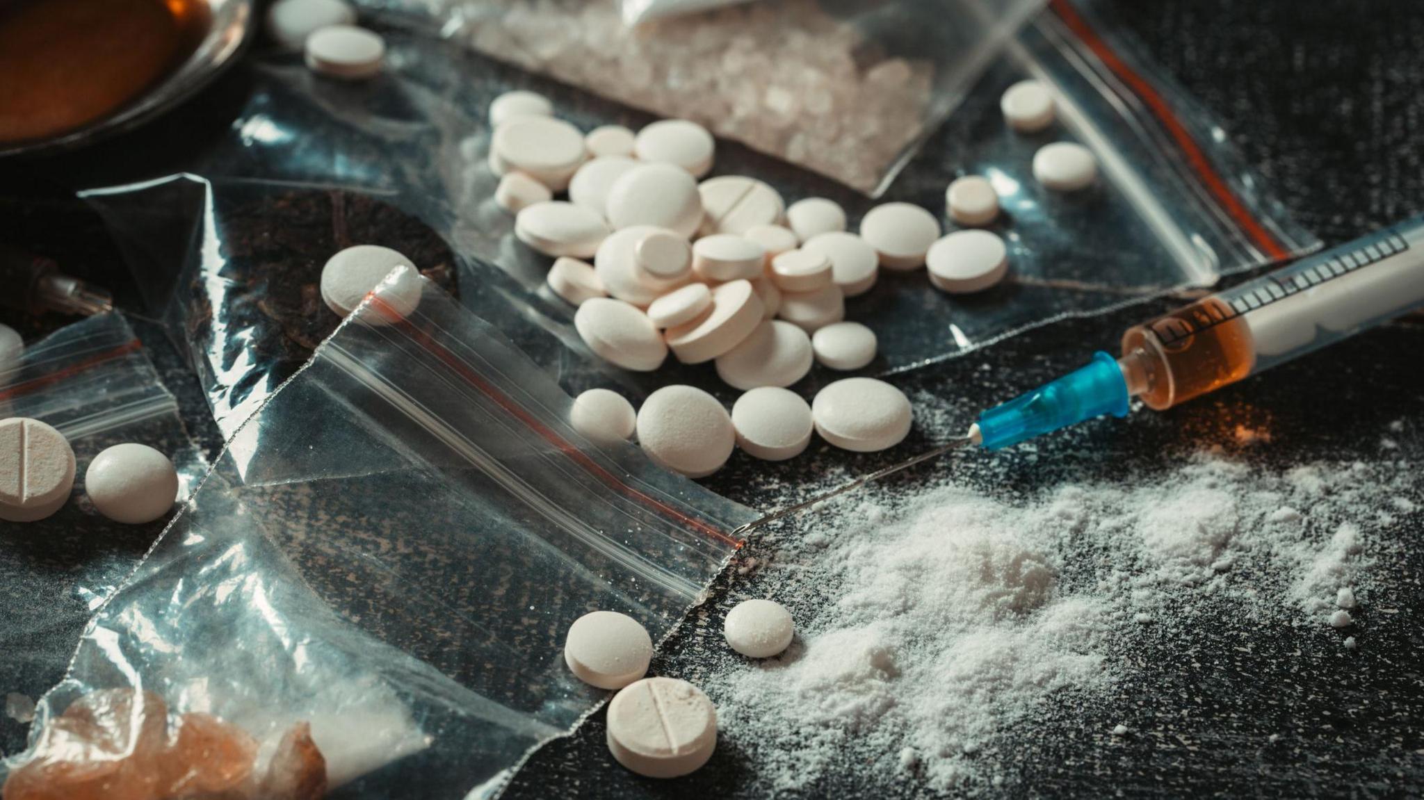 A pile of white pills, white powder, clear plastic bags and a syringe on a dark surface