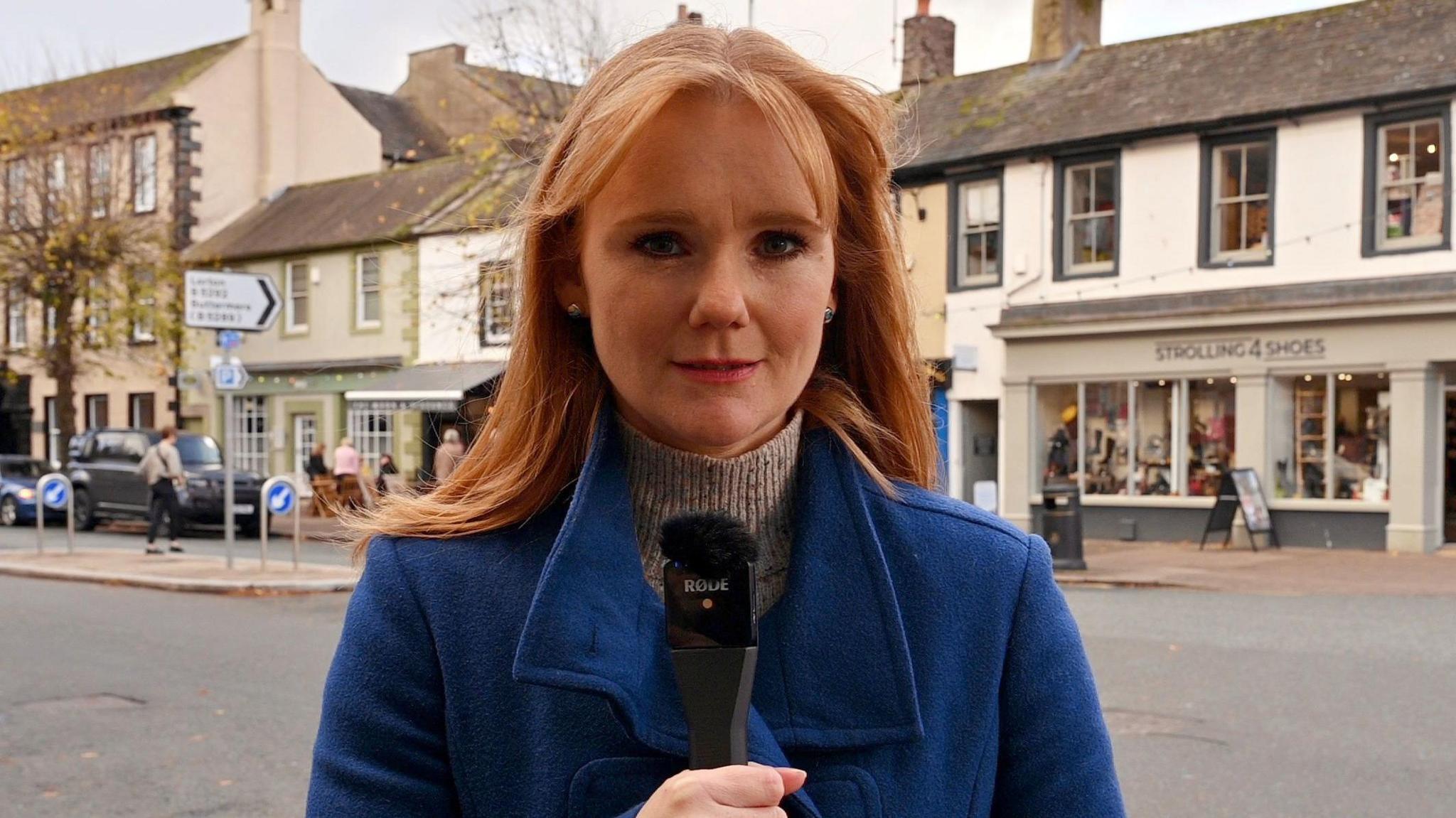 Samantha Down with red hair, wearing a blue coat and holding a microphone, stands on a street. 