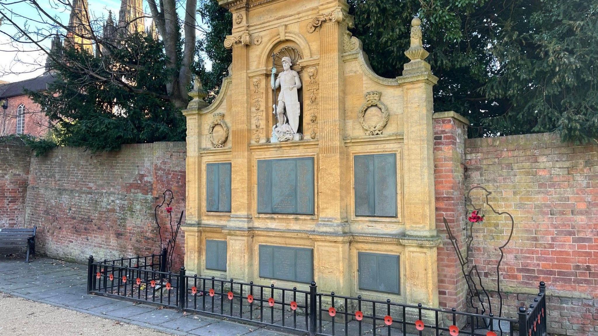 A war memorial featuring a small white statue placed on a structure with names of people who died in the world wars. A black fence with poppies is at the foot of the memorial.