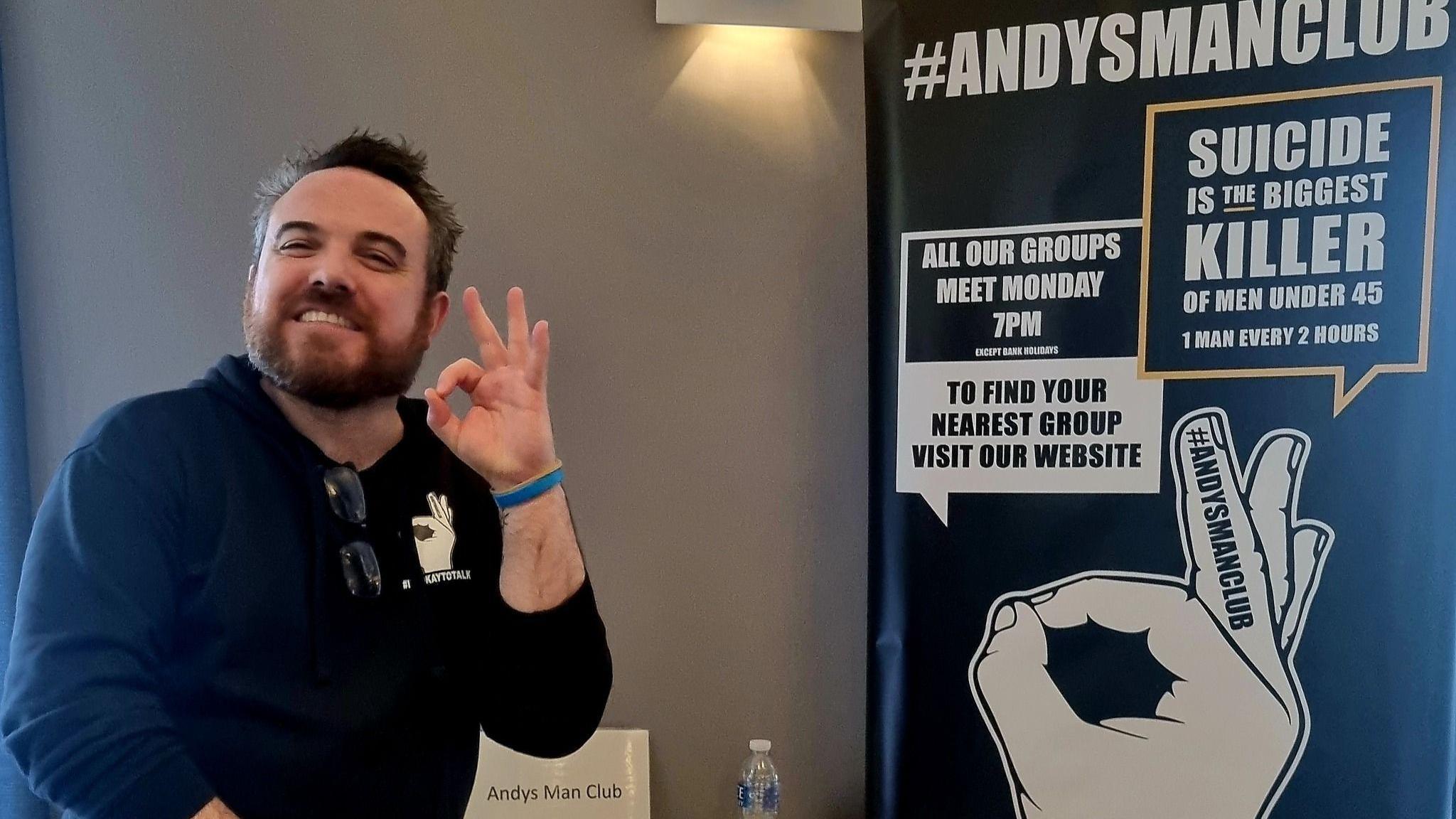 Steve Doyle, from Andysmanclub, is standing to the left holding up one hand and smiling. Next to him is a poster detailing a men's support group that meets every Monday.