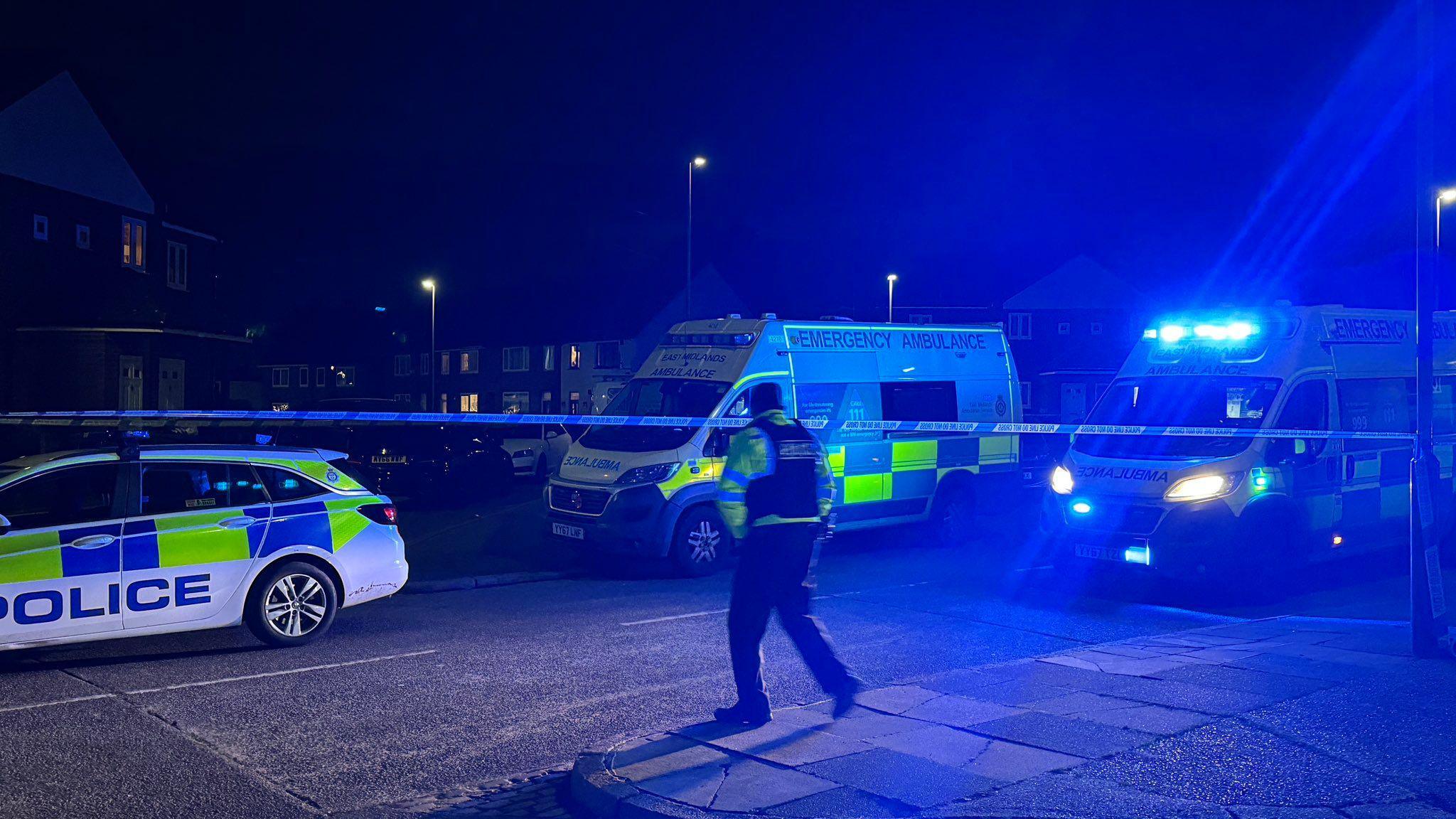 A police cordon in Bloxham Road in New Parks Leicester. A police car and two ambulances are blocking the road. One ambulance has blue lights on. A police officer stands in the middle of the cordon.