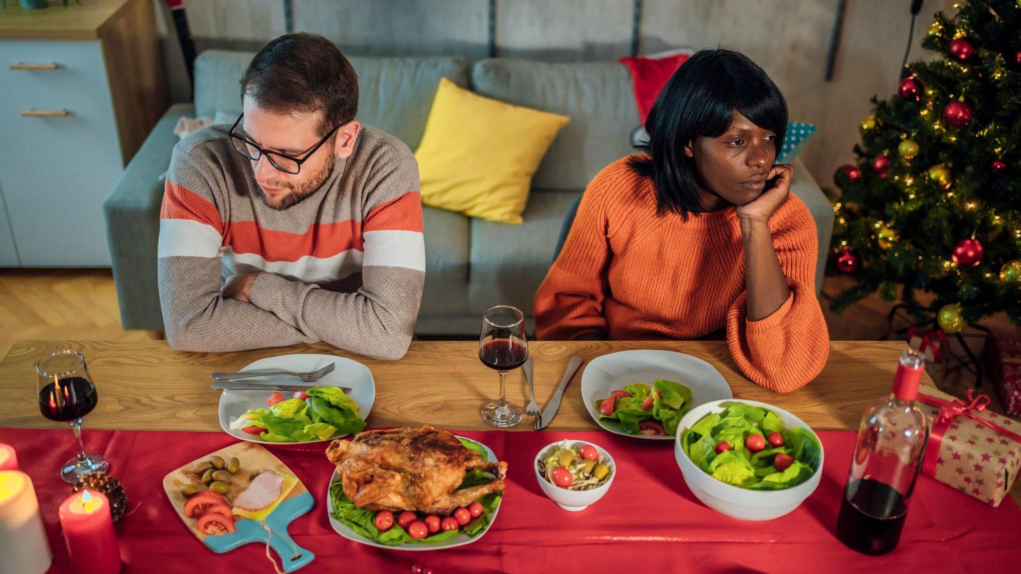 An unhappy couple sit at a festive table ignoring each other. The table has a turkey, lettuce, tomatoes, and wine on the table. There is a sofa behind them and a Christmas tree in the right corner decorated with golden lights and baubles and also red baubles. The man has short brown hair and wears black-rimmed glasses and a brown jumper with two red and white stripes. The woman has black hair and is wearing an orange jumper.