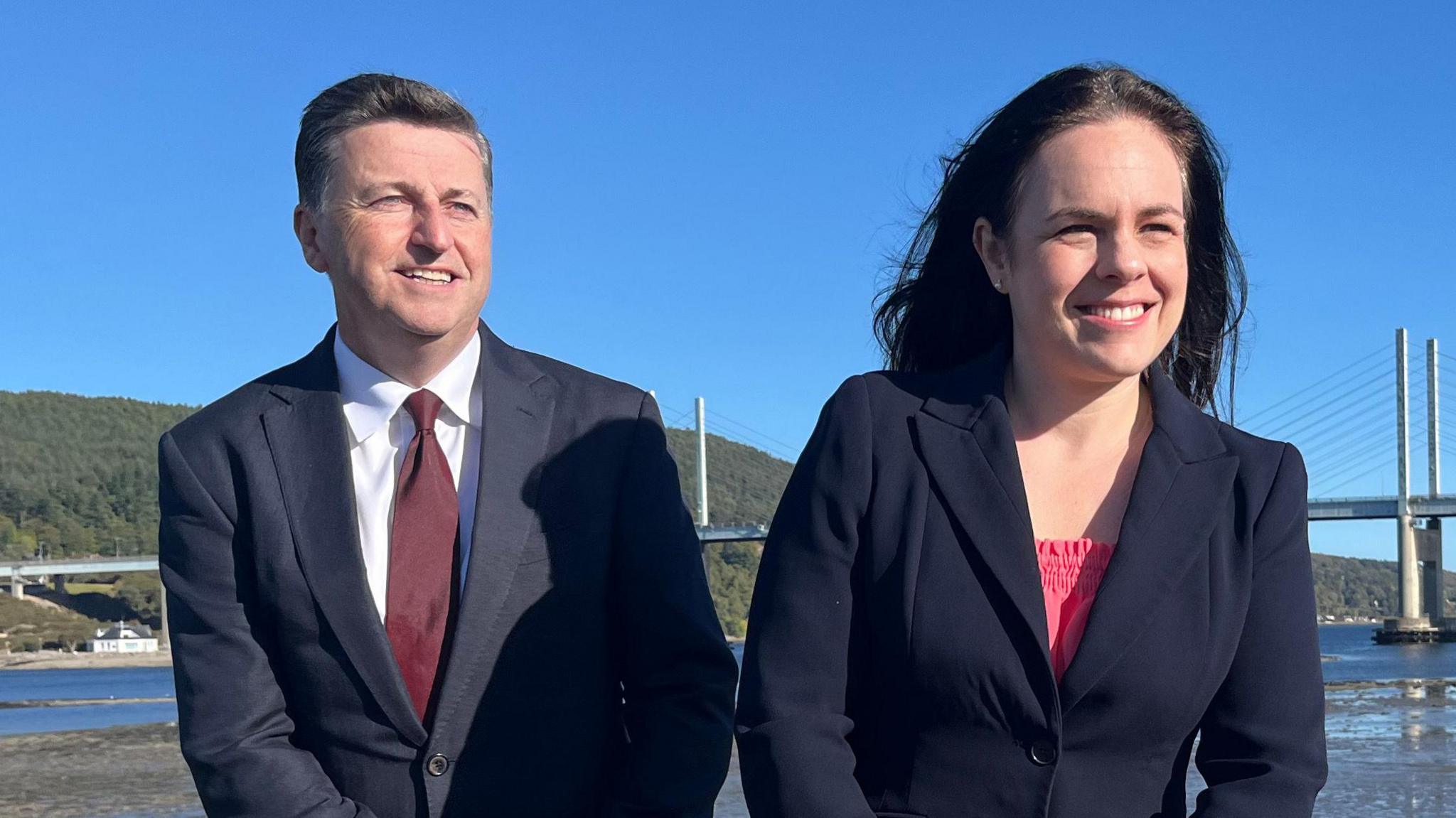 Douglas Alexander, wearing a navy suit and burgundy tie, sitting on a wall next to Kate Forbes who is wearing a dark blazer and pink top. Behind them is a bridge and water.