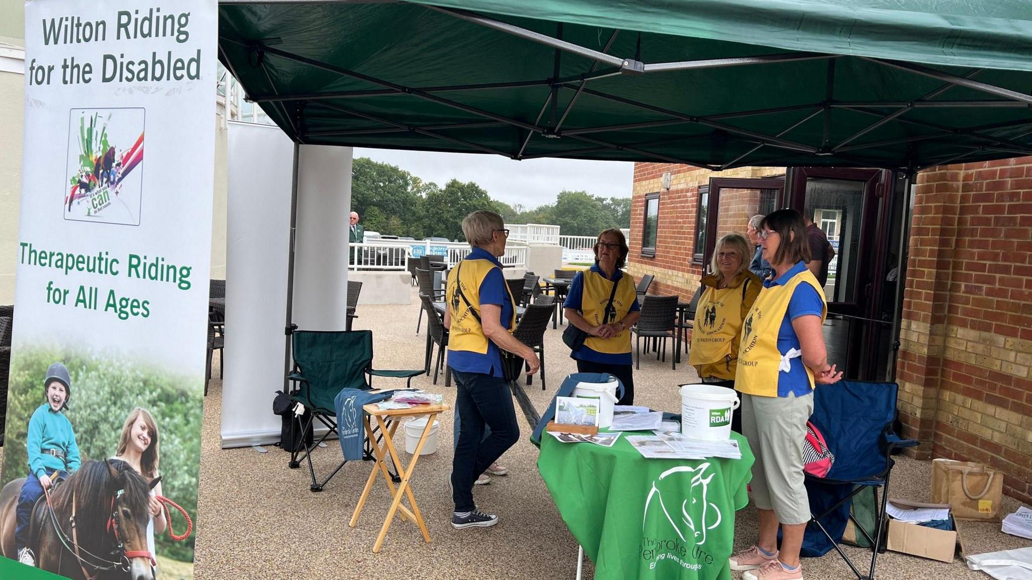 Volunteers in yellow bibs under a gazebo with information and money buckets. There is a large sign that says 'Wilton Riding for the Disabled'.