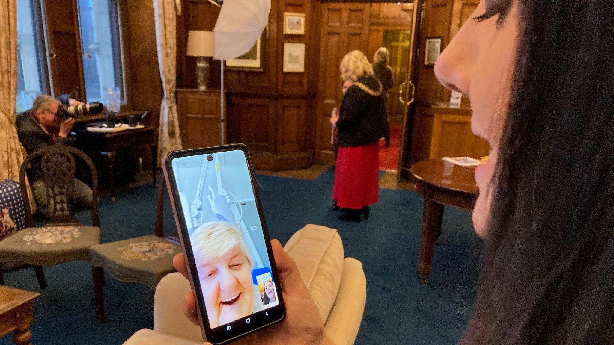 A close-up on a woman's face on a mobile phone being held by another woman with long, dark hair who is standing in a wood-panelled room with a photographer taking pictures in the background of the Lord Mayor. 