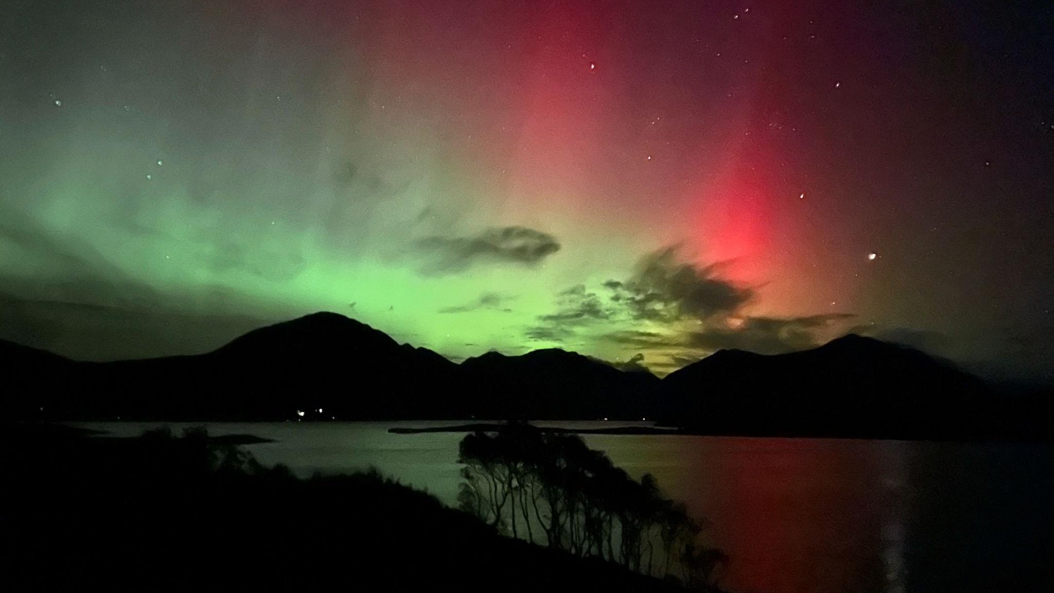 Green and red colours of the aurora over silhouetted mountains with a body of water in the foreground.