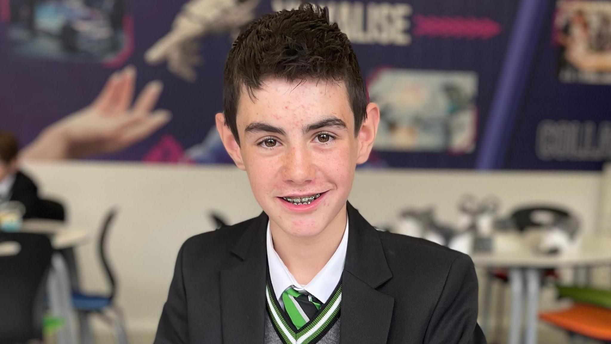 Boy with dark hair wearing black school blazer with green emblem, grey jumper, white shirt and green and black tie